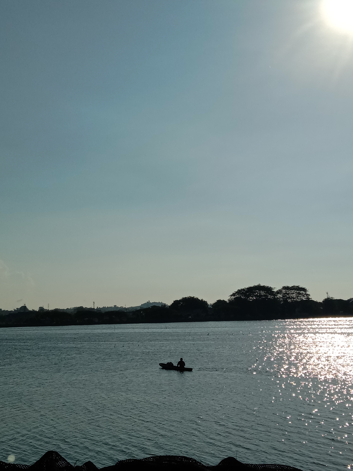 The image depicts a serene waterscape at sunset in Lhokseumawe, Indonesia, likely near the Krueng Geukueh area. The sun, a bright orb with visible rays, is positioned in the upper right corner, casting a warm glow across the scene and creating shimmering reflections on the water's surface. In the foreground, the dark silhouettes of fishing nets or similar structures are visible along the lower edge of the frame. The middle ground is dominated by a vast expanse of calm water, with subtle ripples indicating a gentle breeze. A solitary figure is seated in a small boat, rowing across the water, adding a sense of tranquility and human presence to the otherwise natural setting. The background features a silhouetted treeline, with hints of distant structures and vegetation suggesting the coastline of Lhokseumawe. The sky is a soft gradient of blue, transitioning to a lighter hue near the horizon.