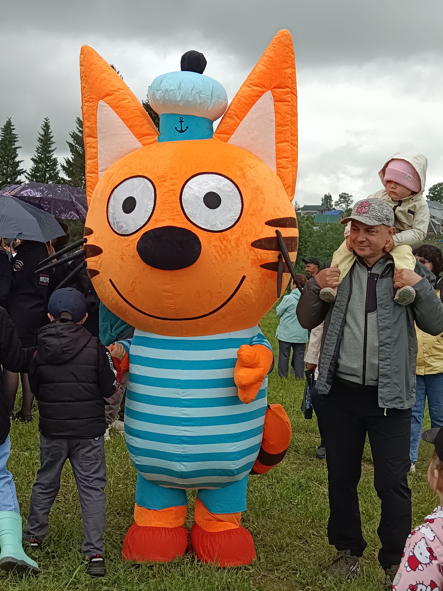 This outdoor image captures a crowded gathering on a grassy field under a heavily overcast sky in Unknown, Unknown. Dominating the foreground is a large, inflated, anthropomorphic orange cat mascot, wearing a light blue and white striped shirt, a light blue sailor hat with a black anchor emblem and a black pom-pom, and red-cuffed orange shoes. The mascot has large white eyes with black pupils, a black nose, a large black smile, and three black "whiskers" on each cheek.
To the right of the mascot, a man with a grey baseball cap and a dark grey jacket carries a young child on his shoulders. The man is smiling slightly, looking towards the right, while the child, wearing a light-colored hooded jacket and a pink knitted beanie pulled low, looks down with a neutral expression.
To the left of the mascot, a young boy with his back to the viewer, wearing a dark blue baseball cap, a black puffer jacket, and dark grey pants, stands close to the mascot, seemingly reaching out with his right hand. Further left and partially obscured, an individual in a dark uniform with "МВД" (MVD) visible on the sleeve holds a black umbrella.
Numerous other adults and children are visible in the mid-ground and background. Many individuals are holding umbrellas of various colors, including black and purple patterned, suggesting rainy or recently rainy conditions. The background features distant evergreen trees and residential buildings under the grey sky. The ground is covered in green grass with some sparse patches.