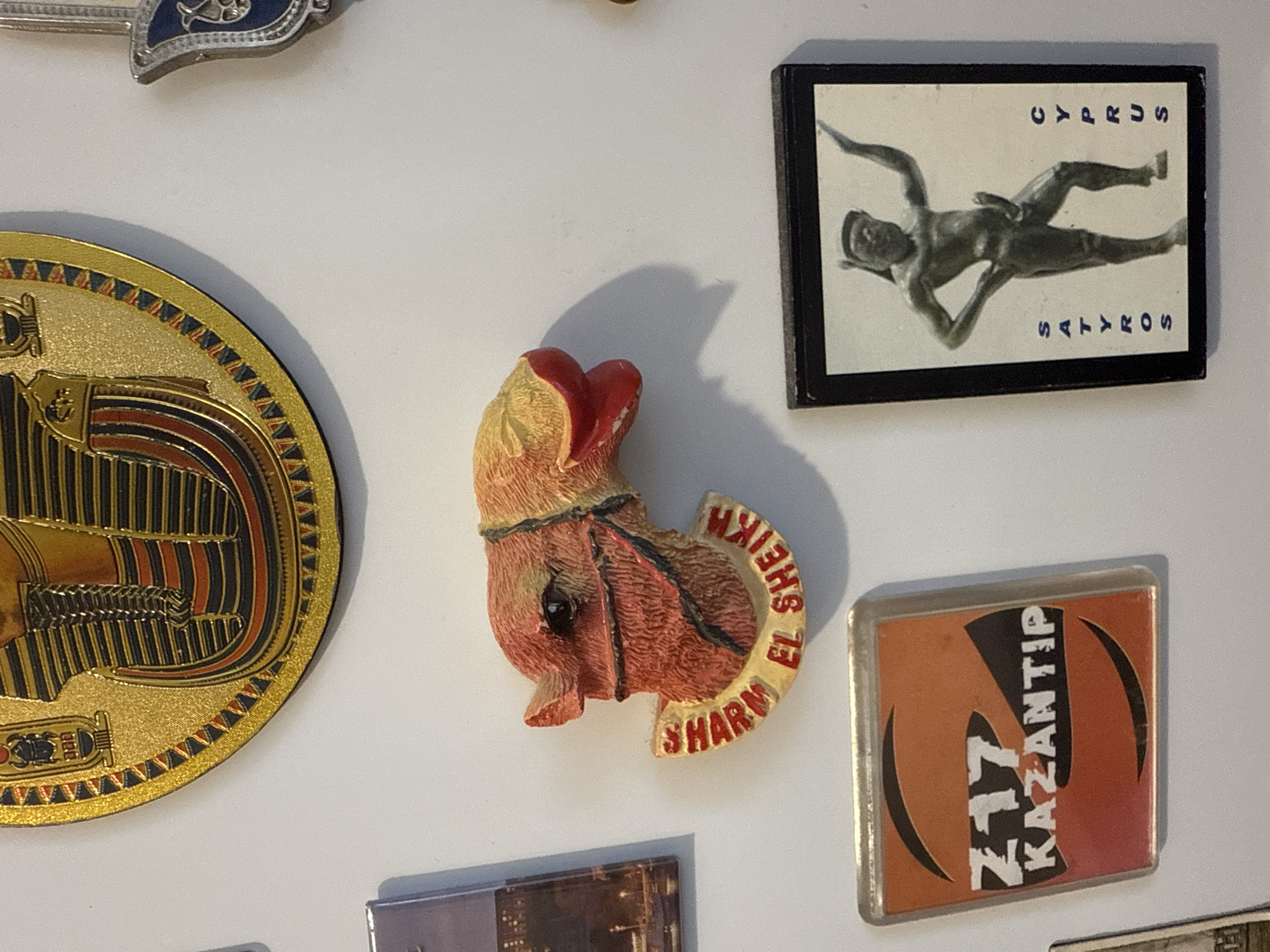 The image displays a collection of magnets on a white surface, likely a refrigerator. From top left to bottom right, the magnets include: a large circular magnet featuring an Egyptian pharaoh's headdress; a camel head magnet with the text "SHARM EL SHEIKH" written on its base; a square magnet with orange and black graphics and the text "217 KAZANTIP"; and a rectangular magnet with a black border, featuring a dark figurine of a male with arms raised and the words "SATYROS" and "CYPRUS" vertically printed on either side. There are also partial views of other magnets on the left and right edges of the frame. The overall scene suggests a domestic setting where souvenirs are displayed. There is no indication of the city of Kyiv, Ukraine, in the image.