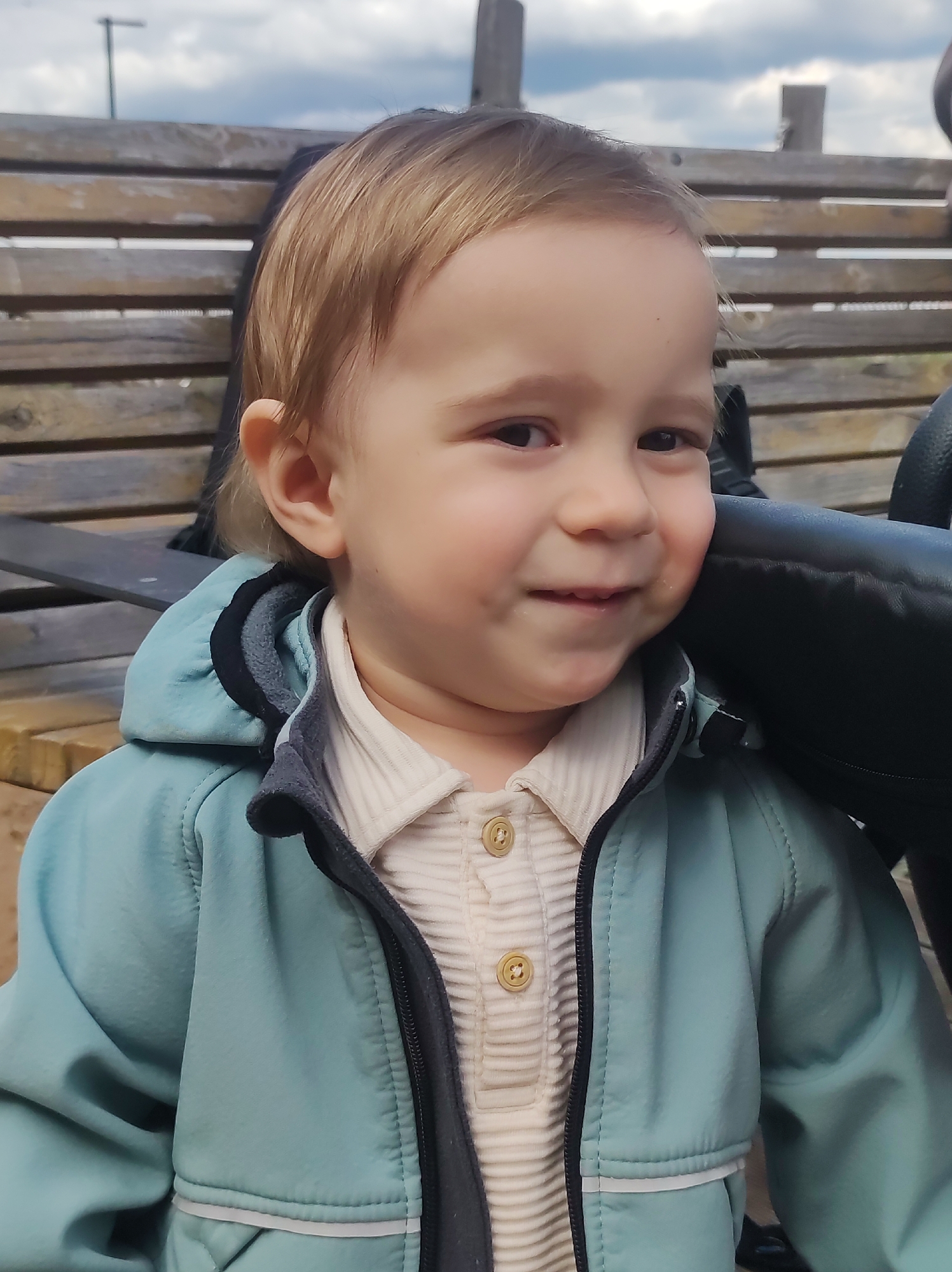 A toddler sits on a wooden bench outside. The child has light brown hair and is wearing a light blue jacket over a cream-colored corduroy shirt with brown buttons. The bench is made of horizontal wooden slats. Behind the child is a black object, possibly a stroller wheel. The sky is visible in the background. It is not possible to verify the location in the city of Chaikovsky, Russia, based solely on the visual information.