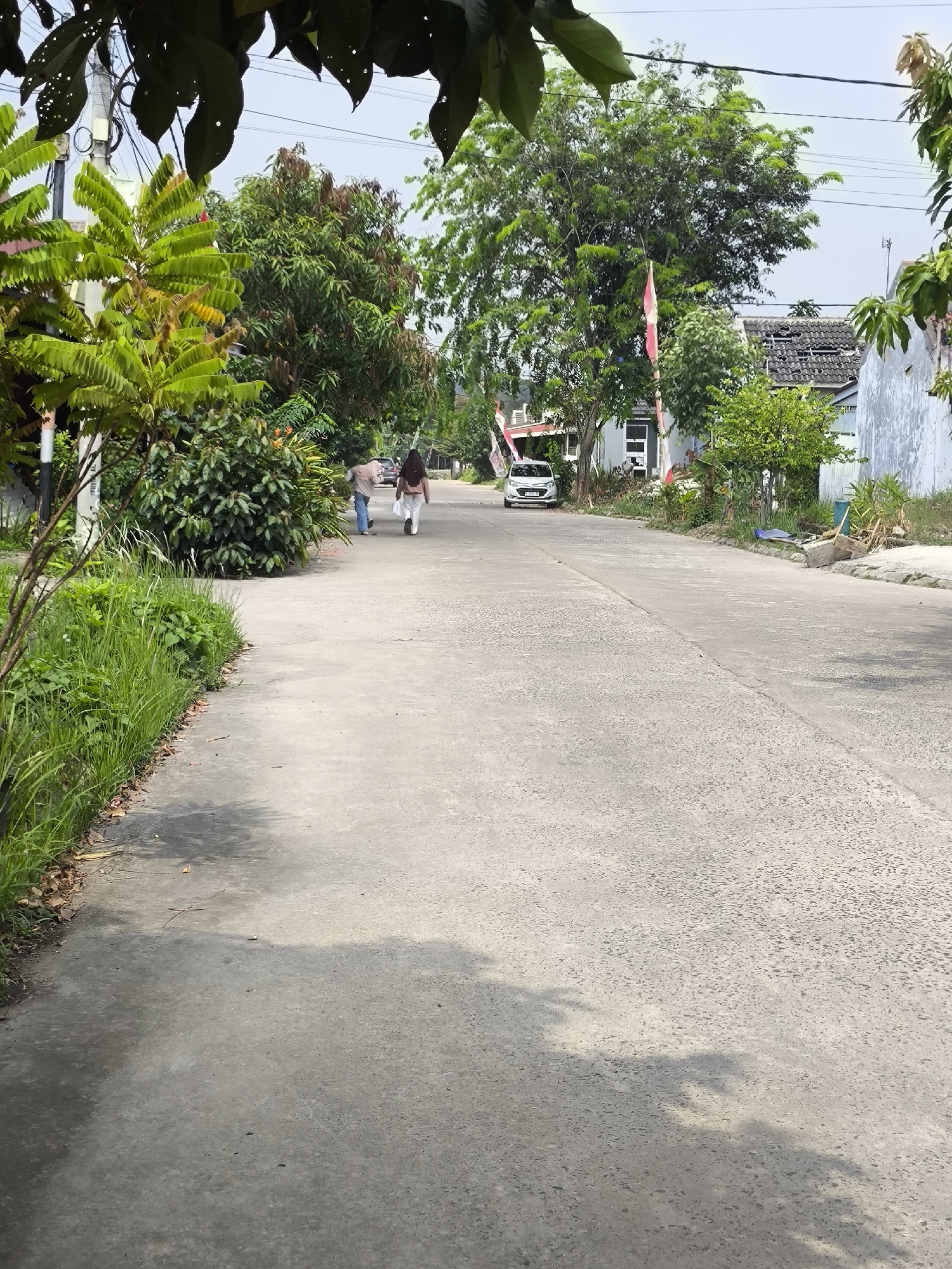 The image depicts a residential street in Cilegon, Indonesia, likely a suburban area given the presence of houses, trees, and a paved road. Two individuals are walking away from the camera on the right side of the road. The person in front is wearing white pants and a light-colored top, while the person behind is wearing a dark hijab and white pants. A white car is parked further down the road on the left. The street is flanked by houses and lush greenery, with trees and bushes lining the sides. There are also power lines visible above the trees. The overall impression is of a typical day in a quiet Indonesian neighborhood.