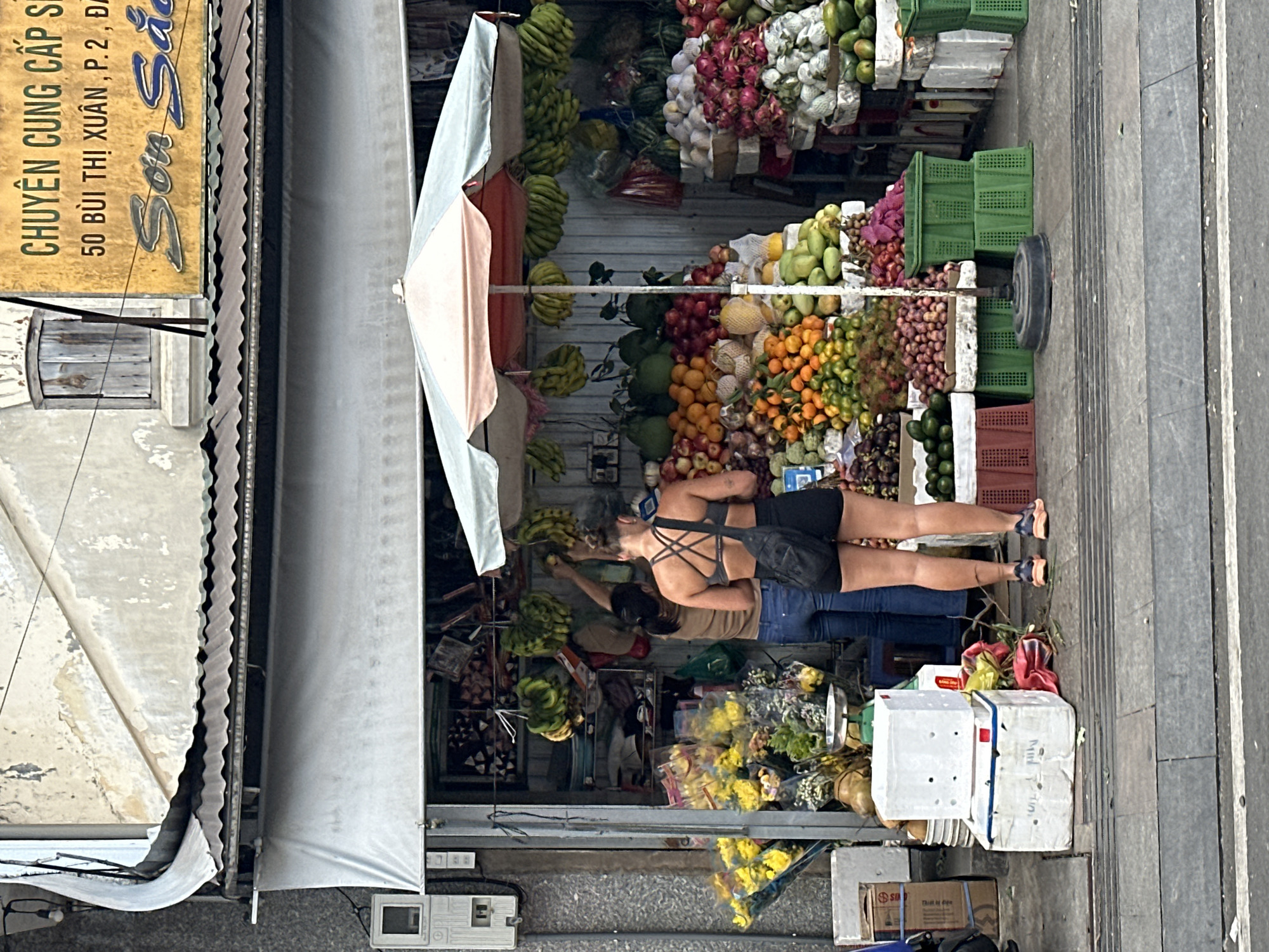 The image depicts an outdoor fruit stand in Phường Xuân Hương - Đà Lạt, Vietnam. Two women are present. One woman, wearing blue jeans and a tan shirt, is standing behind the fruit stand, appearing to be interacting with the fruit. The other woman, wearing black shorts and a black athletic top, stands in front of the fruit stand, looking at the merchandise. The stand is laden with a variety of fresh fruits, including bananas, oranges, apples, dragon fruit, and what appear to be pomelos and mangoes. Several green plastic crates are used to display the produce. Yellow flowers are arranged in bouquets to the left of the fruit stand. The background shows a building with a sign in Vietnamese, indicating the establishment's name and address. A white awning covers part of the stand. The scene is set on a paved sidewalk, with a road visible in the foreground.