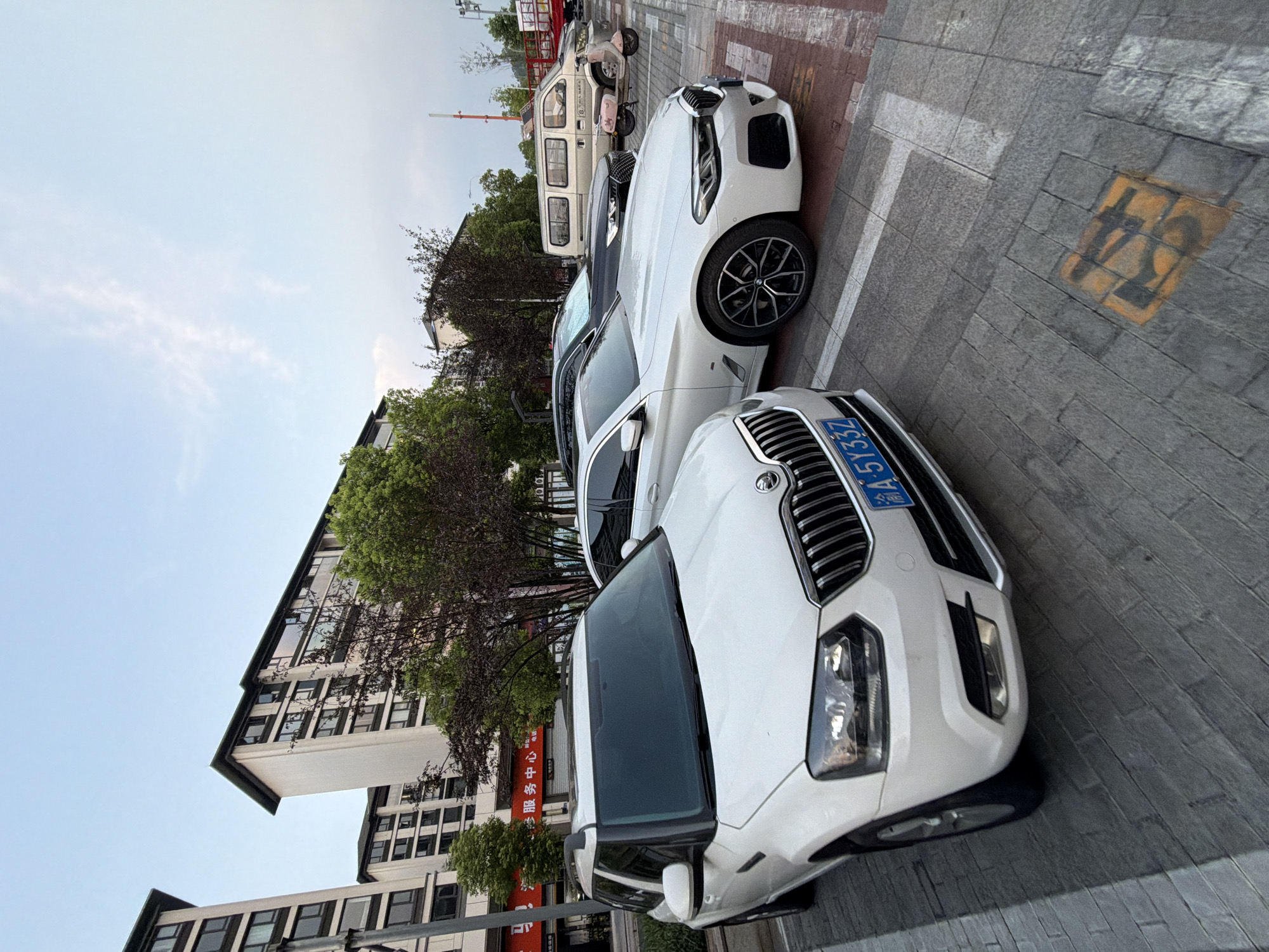 The image shows a parking lot scene in the city of Jiangjin, China. In the foreground, a white Skoda Yeti SUV is parked, facing left. The vehicle has a license plate that reads "A 5Y33Z". To its right, a white BMW sedan is parked, partially obscured by the Skoda. Behind these two cars, a black sedan is parked, and further back, a silver van is visible. A pink scooter is parked to the right of the silver van. The background features apartment buildings and trees. The ground is paved with gray brickwork, and parking lines are visible. The sky is partly cloudy, with a soft, diffused light suggesting it's either early morning or late afternoon.