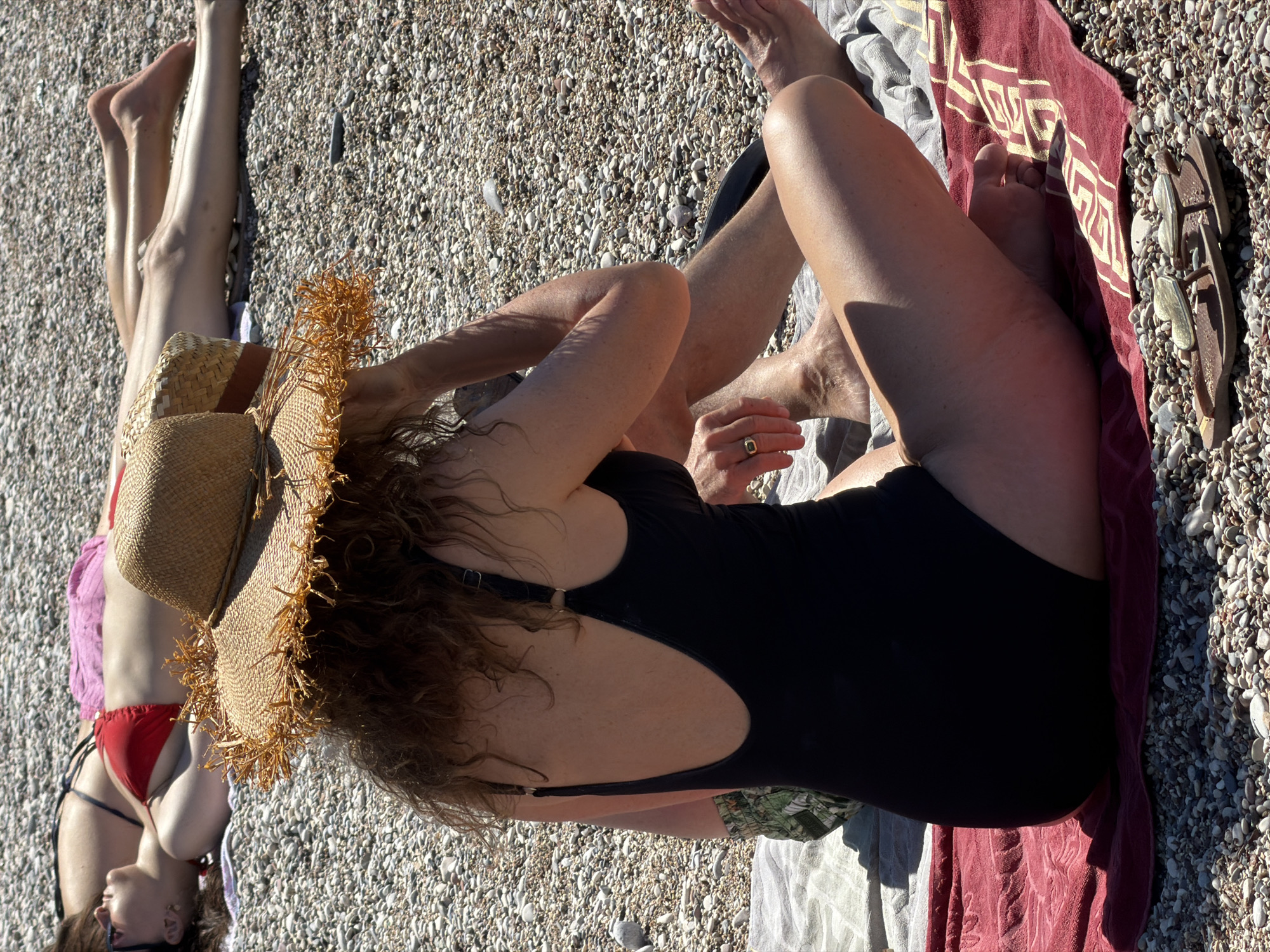 The image depicts a beach scene in Bečići, Montenegro. Two individuals are present. One is lying on their back, wearing a red bikini and a straw hat partially obscures their face. The other, wearing a black one-piece swimsuit, is seated with their legs crossed and arms raised. They are also wearing a straw hat. Both are on towels on the pebbled beach. A maroon towel and a pair of sandals are visible nearby. The ground is a pebble beach, typical for the area. The scene is brightly lit, suggesting daytime.