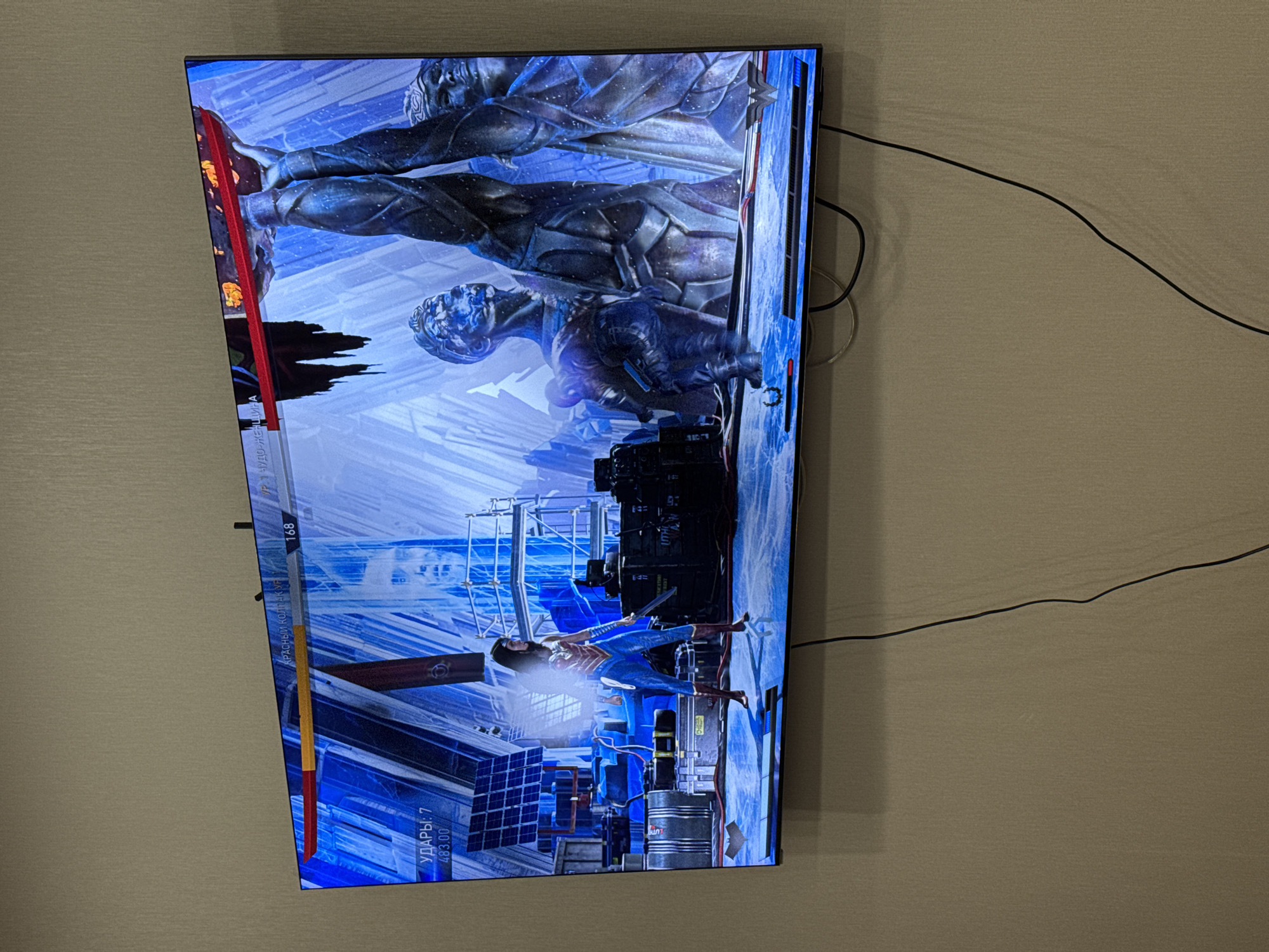 The image shows a television screen displaying a scene from what appears to be a video game. The game scene depicts a female character in blue and red attire, holding a sword, and standing on what appears to be a rooftop or platform in an urban environment. The background features high-rise structures, scaffolding, and industrial equipment. Above her are three large, statuesque figures appearing in a silvery-blue tone. The text on the screen is in a non-English language, possibly Russian, and includes game-related information like a health bar. The television is mounted on a wall, and several black cables are visible adjacent to it. The environment appears to be an indoor setting, with a textured wall in the background. The scene is depicted in shades of blue, gray, red, and black.