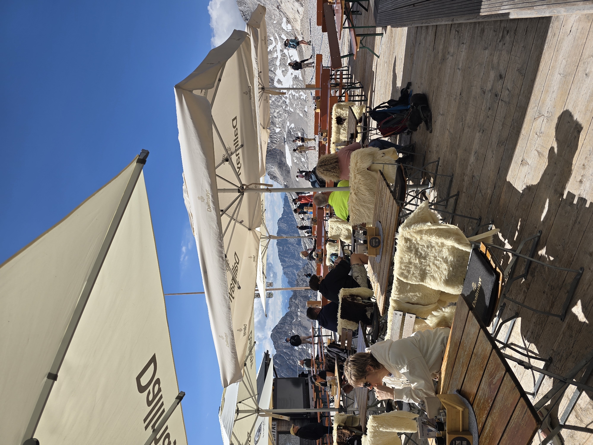 This is an outdoor photograph taken on a sunny day at a mountain restaurant, likely at the Zugspitze, Germany's highest peak, near Garmisch-Partenkirchen. The scene is set on a wooden deck with several tables and chairs, some covered by large white umbrellas that bear the "Dallmayr" logo.
In the foreground, a woman with short blonde hair and sunglasses is seated at a wooden table, looking to her left. She is wearing a white hooded sweatshirt. The table in front of her has a wooden crate with salt and pepper shakers and a small menu. Her chair is draped with a fluffy white sheepskin.
Further back, other patrons are seated at tables, some eating or talking. One person in a bright green jacket is visible. Several chairs are also covered with sheepskins, suggesting a cooler mountain climate. In the background, a stunning mountain landscape with snow-capped peaks under a clear blue sky dominates the view. People can be seen walking on a snowy path in the distance, and some buildings are visible on the mountainside. Shadows of the umbrellas and people are cast on the wooden deck, indicating the time of day is likely mid-day. A black dog is lying on the wooden deck in the lower right of the frame.