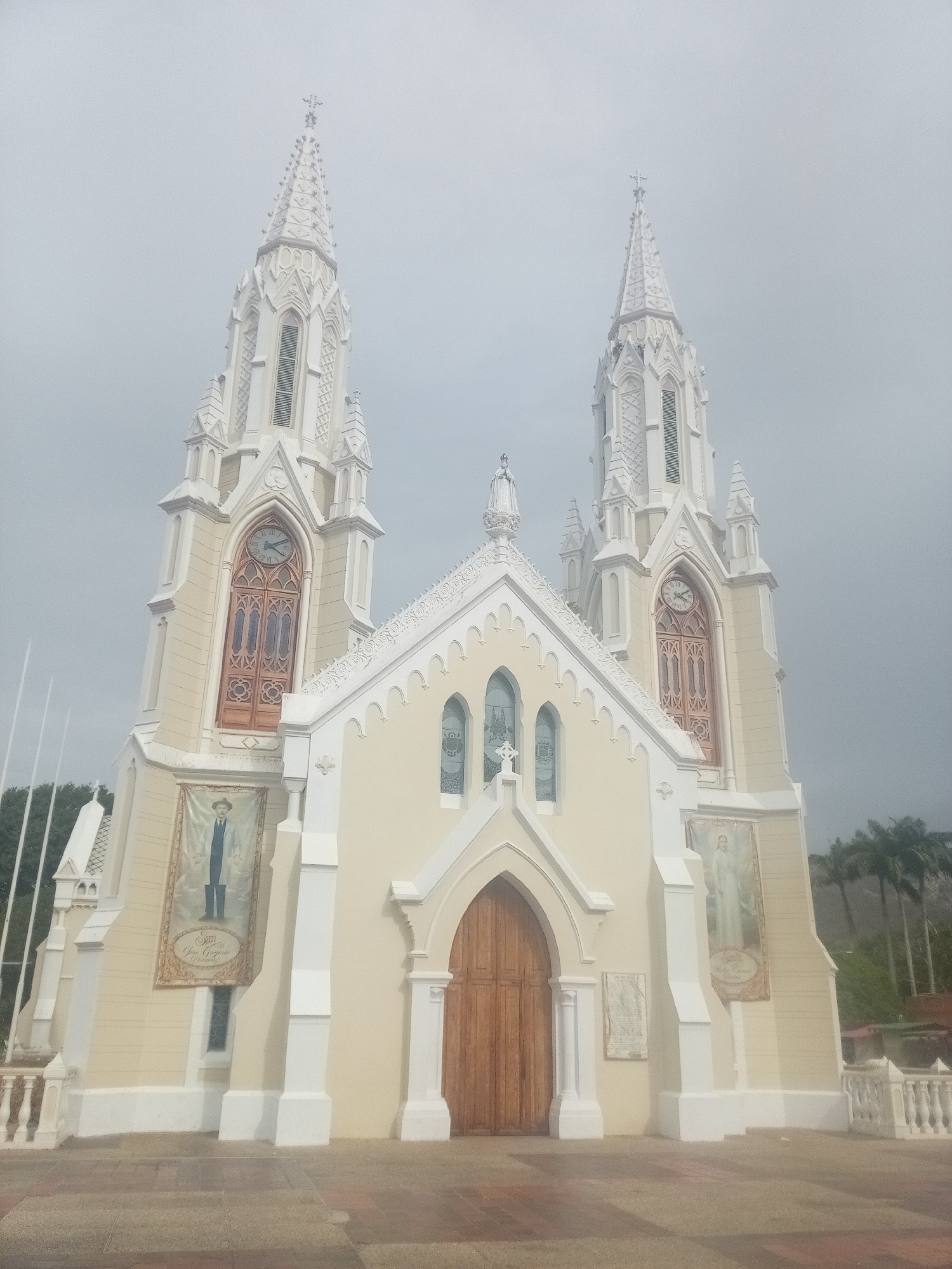 The image depicts the Basilica of Our Lady of El Valle, located in El Valle del Espíritu Santo, Venezuela. The basilica is a large, ornate, neo-Gothic structure with two tall spires. Each spire contains a clock face. The basilica has a pale yellow facade with white trim. Banners hang on the sides of the building. A large wooden door is centered on the facade. The sky is overcast. Palm trees and other vegetation can be seen in the background. A paved area is in front of the basilica.