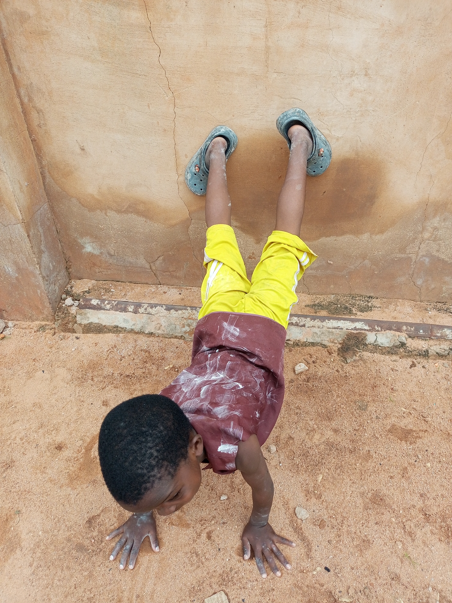 A young child is performing a handstand against a textured, pale orange wall. The child is facing away from the viewer, with their head tilted down and their hands on the sandy ground. They are wearing a burgundy shirt with white splotches and bright yellow shorts. Blue Crocs are on their feet, which are against the wall. The environment appears to be outdoors, with a dusty ground and a cracked wall. The overall impression is one of youthful energy and playfulness in a simple, outdoor setting. The location is a street in Tempure, Nigeria.