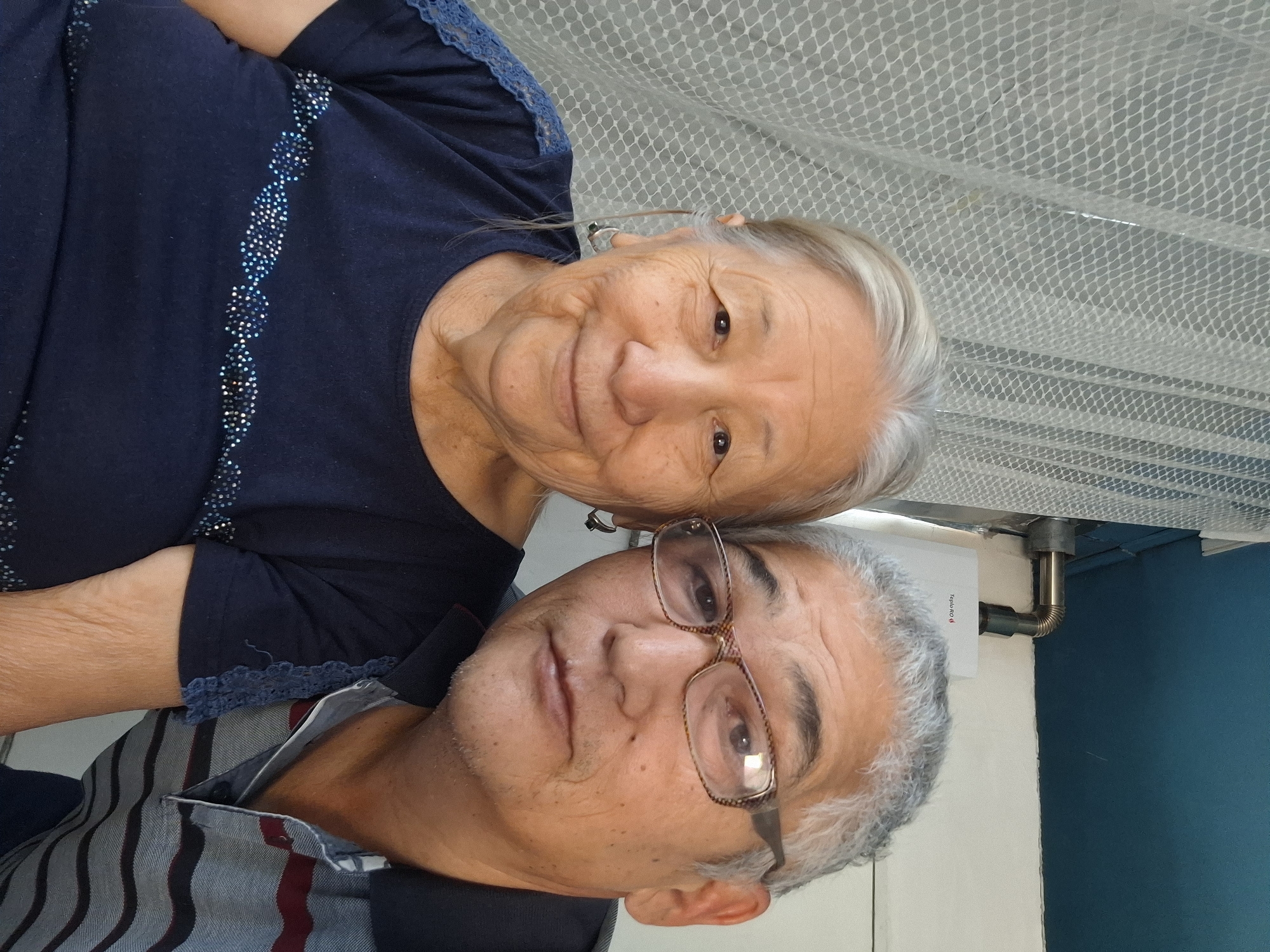A selfie shows an elderly woman with graying hair and a man with graying hair and glasses. The woman is wearing a dark blue shirt adorned with a beaded pattern. The man is wearing a striped polo shirt. Behind them, a white net curtain is visible to the left, and a white water heater with a metal pipe is mounted on the wall to the right. The environment appears to be indoors. The photo is taken in Almaty, Kazakhstan.