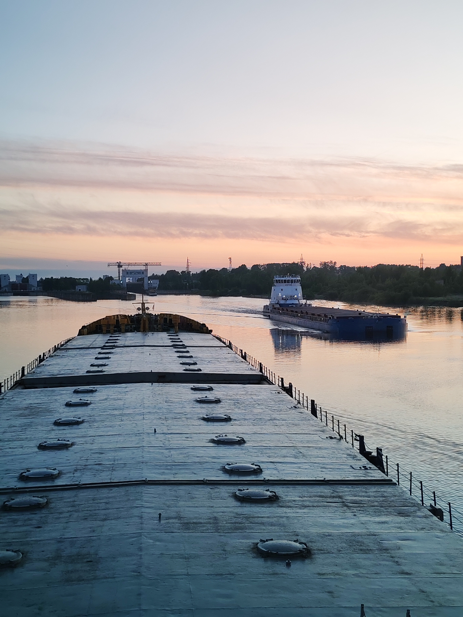 The image shows a wide river with two barges during sunset or sunrise. The foreground barge, presumably where the photo was taken from, has a flat metal deck with rows of hatches. A black railing lines the right edge of the deck.
In the middle ground, another barge moves on the river, displaying a blue hull and a white superstructure. On the riverbanks, are trees, buildings, powerlines, and a crane.
The sky above is a gradient of blue to pale pink with faint wisps of clouds. The water is calm and reflects the colors of the sky.