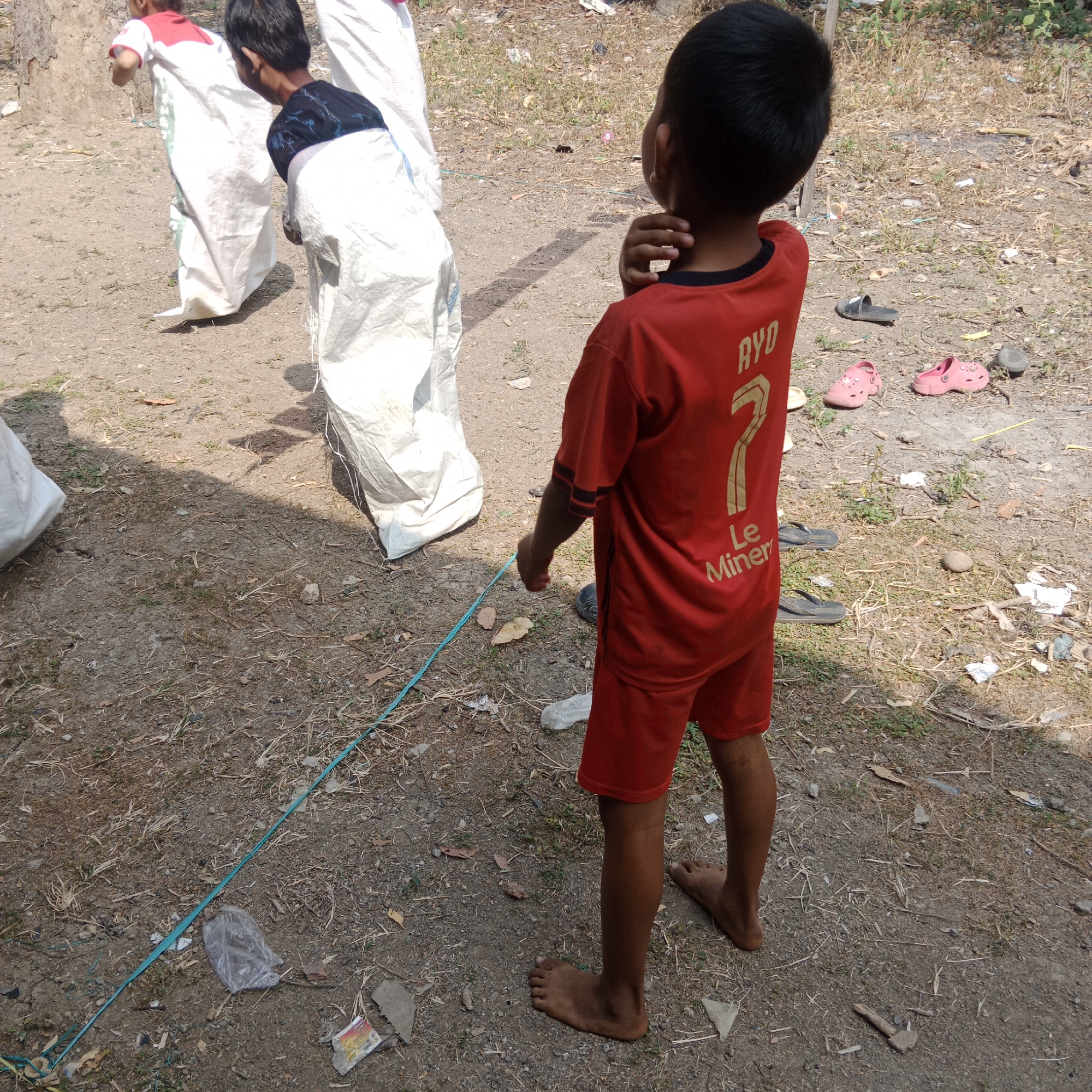 In Tegowanu Kulon, Indonesia, two children participate in a sack race. One child, in the foreground, wears a red t-shirt with a "7" and text that reads "RYD Le Miner." They stand barefoot, facing away from the camera, with their hand to their neck. Behind them, another child is halfway inside a white sack, their legs visible as they hop. A third child, also in a sack, is further in the background. The ground is dry and dusty, with sparse dry grass and scattered debris, including some plastic bags and a blue string marking a race lane. Several pairs of children's footwear, including pink sandals and dark flip-flops, are scattered on the ground. The overall scene depicts an outdoor activity in a rural or semi-rural setting.