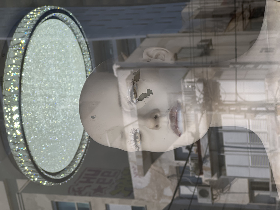 The image displays a head-and-torso view of a female mannequin, positioned behind a reflective glass surface in Tel-Aviv, Israel. The mannequin has a pale complexion, prominent white eyelashes, light-colored eyes, and reddish-brown lips with some white discoloration. Distinct damage includes a dark circular mark on its forehead and a fragmented crack below its left eye. Reflected on the glass above the mannequin is a large, circular, crystal-studded light fixture. The external urban environment, visible through and reflected on the glass, comprises an older, weathered multi-story building to the left, which exhibits graffiti (including the letters "CHU", a star, and "EE" in red and black), a blue-framed window with a white roller shutter, and an external air conditioning unit. To the right, a taller, modern building with a vertical architectural pattern is present. Numerous overhead utility wires traverse the mid-ground and lower portions of the scene.