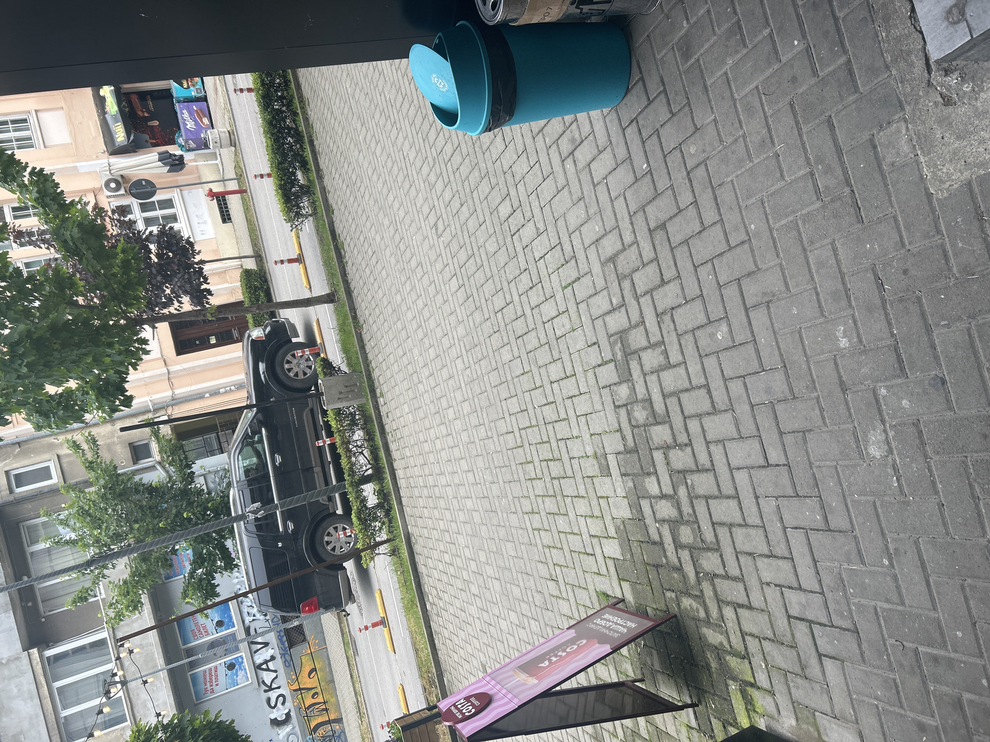 The image, taken from within a structure, shows an external street scene in Varna, Bulgaria. The street is paved with gray bricks laid in a herringbone pattern. A blue cylindrical trash can with a lid is near the upper left. A black SUV is parked parallel to the sidewalk, and there is a tree beside the vehicle. On the left of the frame, there are buildings with windows and foliage. Street lights extend across the frame from the buildings. A Costa Coffee advertising sandwich board is positioned in the lower left corner of the frame. The sidewalk is a light gray color, and the street has several yellow cones near the vehicle.