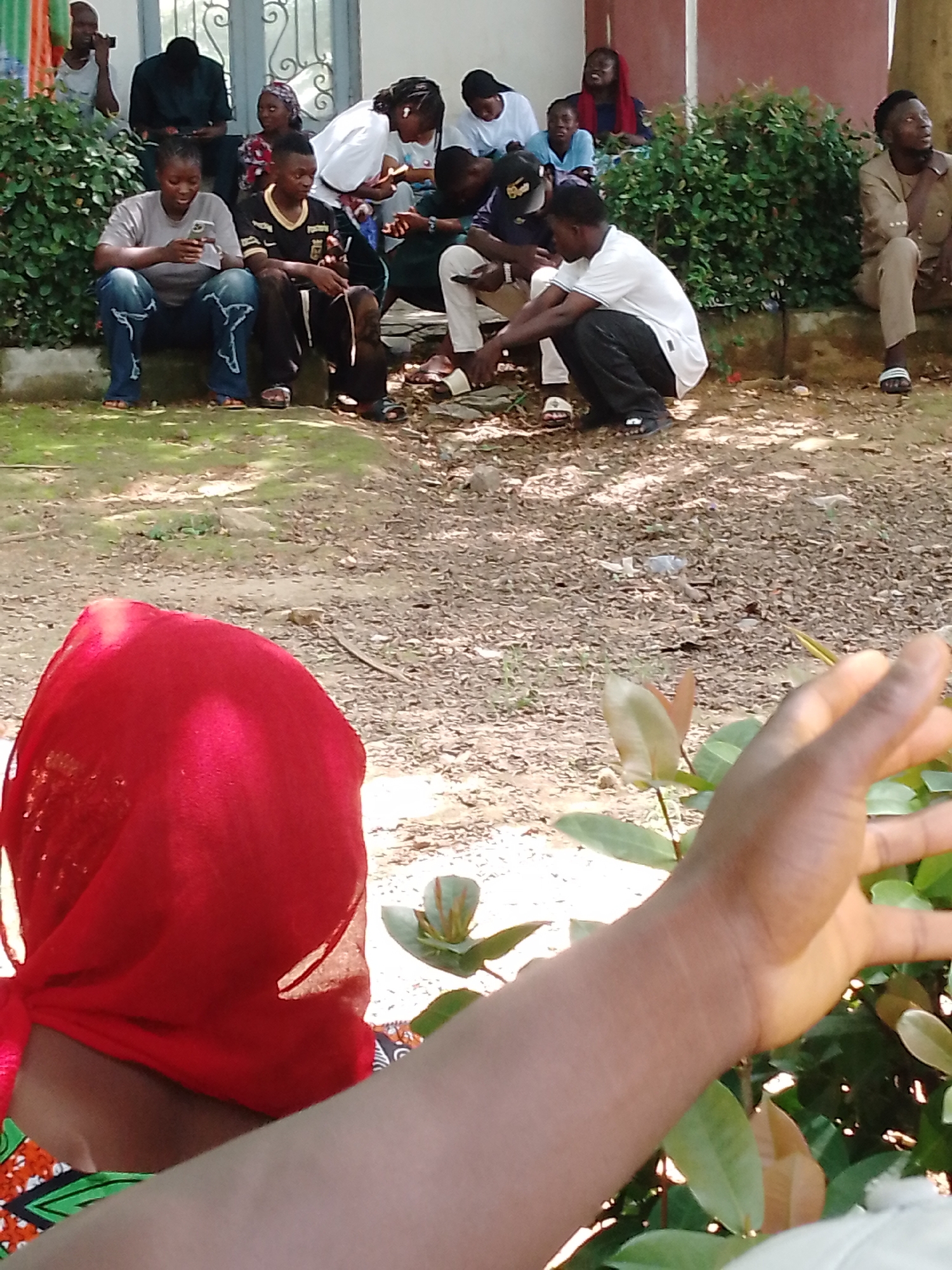 The image depicts a group of people, seemingly young adults, gathered outdoors. Some are seated on a low wall or steps, while others are standing. Several individuals are looking at or interacting with their mobile phones. The environment appears to be a somewhat unkempt area with dry leaves and dirt on the ground, and lush green bushes in the background. In the foreground, a portion of a person's arm and hand is visible, along with a bright red head covering and part of a colorful garment. The overall atmosphere suggests a casual, informal gathering, possibly in a residential or public outdoor space in Akwanga, Nigeria.