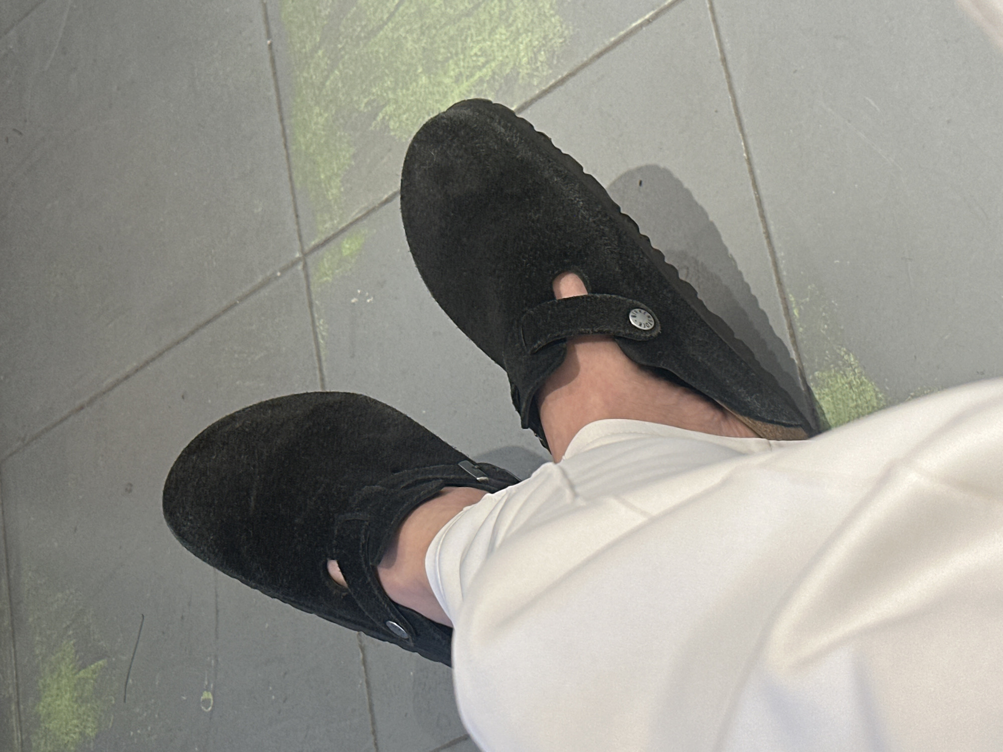 A first-person perspective image shows a pair of black suede Birkenstock clogs on a person's feet, wearing white pants. The clogs are positioned on a gray tiled floor, with faint green markings visible on some tiles, possibly moss or paint. The lighting suggests an indoor or shaded outdoor environment. The scene is consistent with an urban setting.