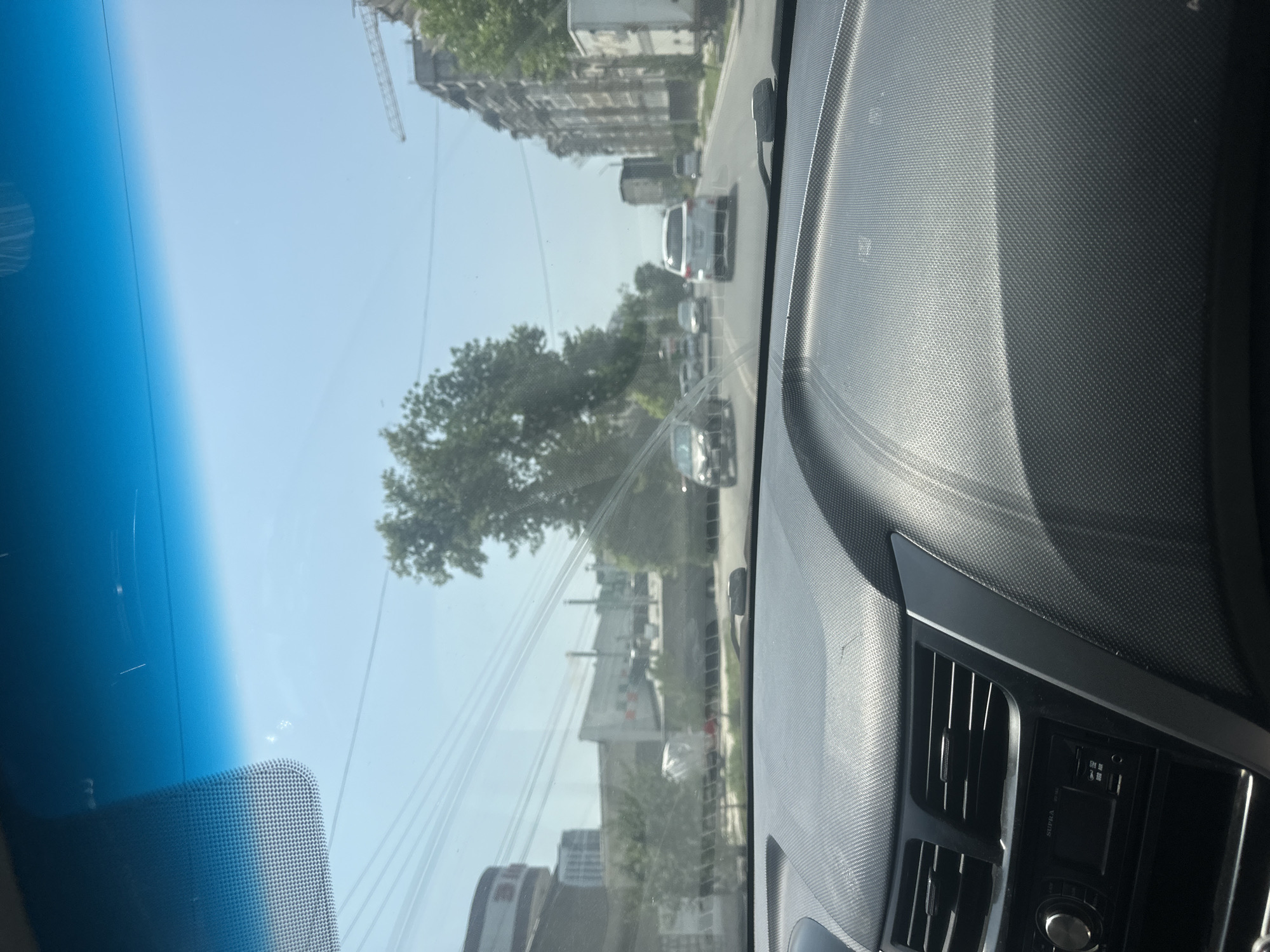 The image is taken from the interior of a vehicle, looking forward through the windshield. The dashboard, with its textured surface and air vents, occupies the foreground. Outside, the view is of a city street on a clear, sunny day.
There are several cars on the road, moving in the same direction. Some buildings are visible on either side of the street, including what appears to be an apartment building on the right and other structures on the left. There are also trees lining the street, and power lines are visible crisscrossing the sky. A construction crane is also present in the distance. The overall scene suggests an urban environment. The specific location is Manas, Kyrgyzstan.
