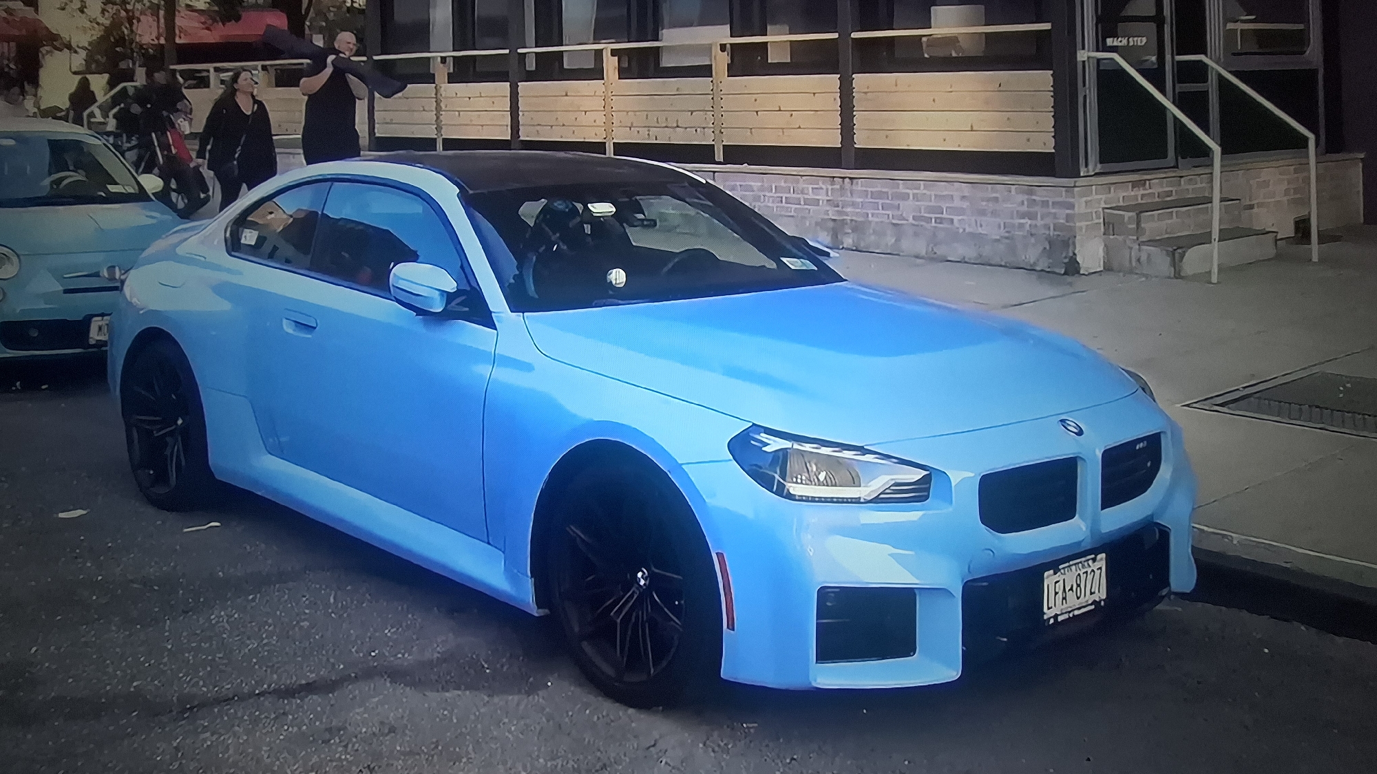 The image shows a light blue BMW M2 coupe parked on a street. The car has a black roof and black wheels. A license plate that reads "LFA-8727" is visible. In the background, a light blue Fiat 500 is also parked. People can be seen walking behind the cars. A building with a light-colored wooden facade and a brick base is present in the background. The location appears to be an urban environment, likely a city street.