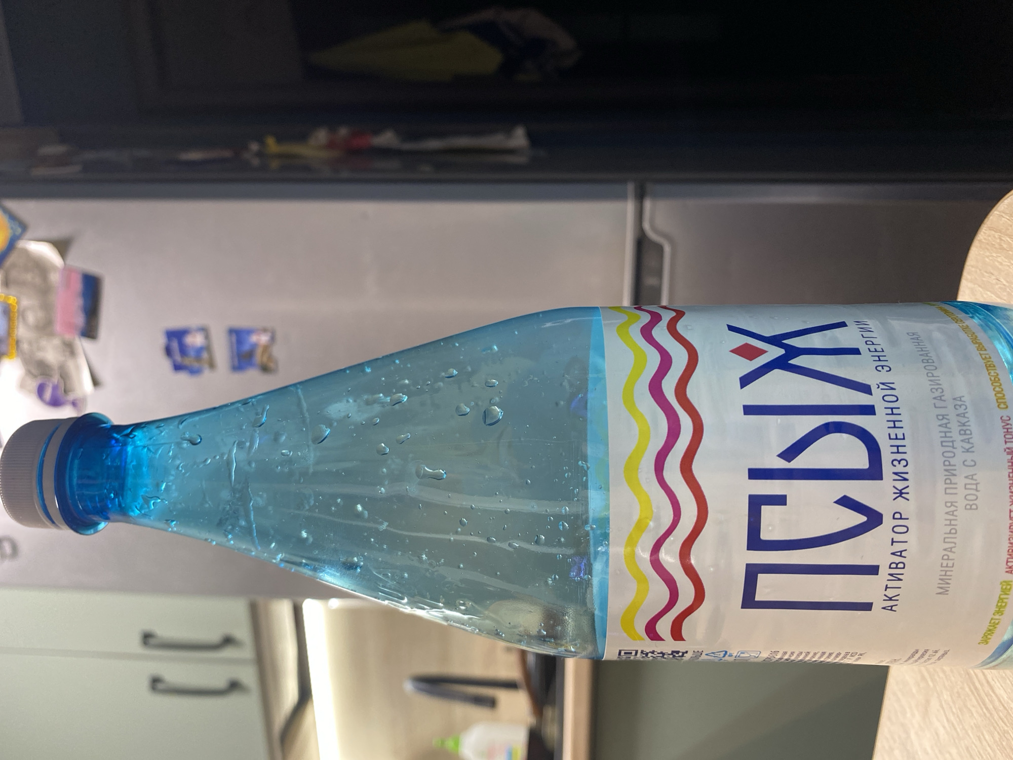 A close-up shot focuses on a blue water bottle, labeled "ПСЫЖ" in large, bold letters, along with other Cyrillic text. The bottle is covered in water droplets, suggesting it's been refrigerated or recently taken out of a cold environment. The background shows a kitchen setting, including a refrigerator with magnets and items on the shelves. The bottle is placed on a light brown surface, possibly a countertop or table.