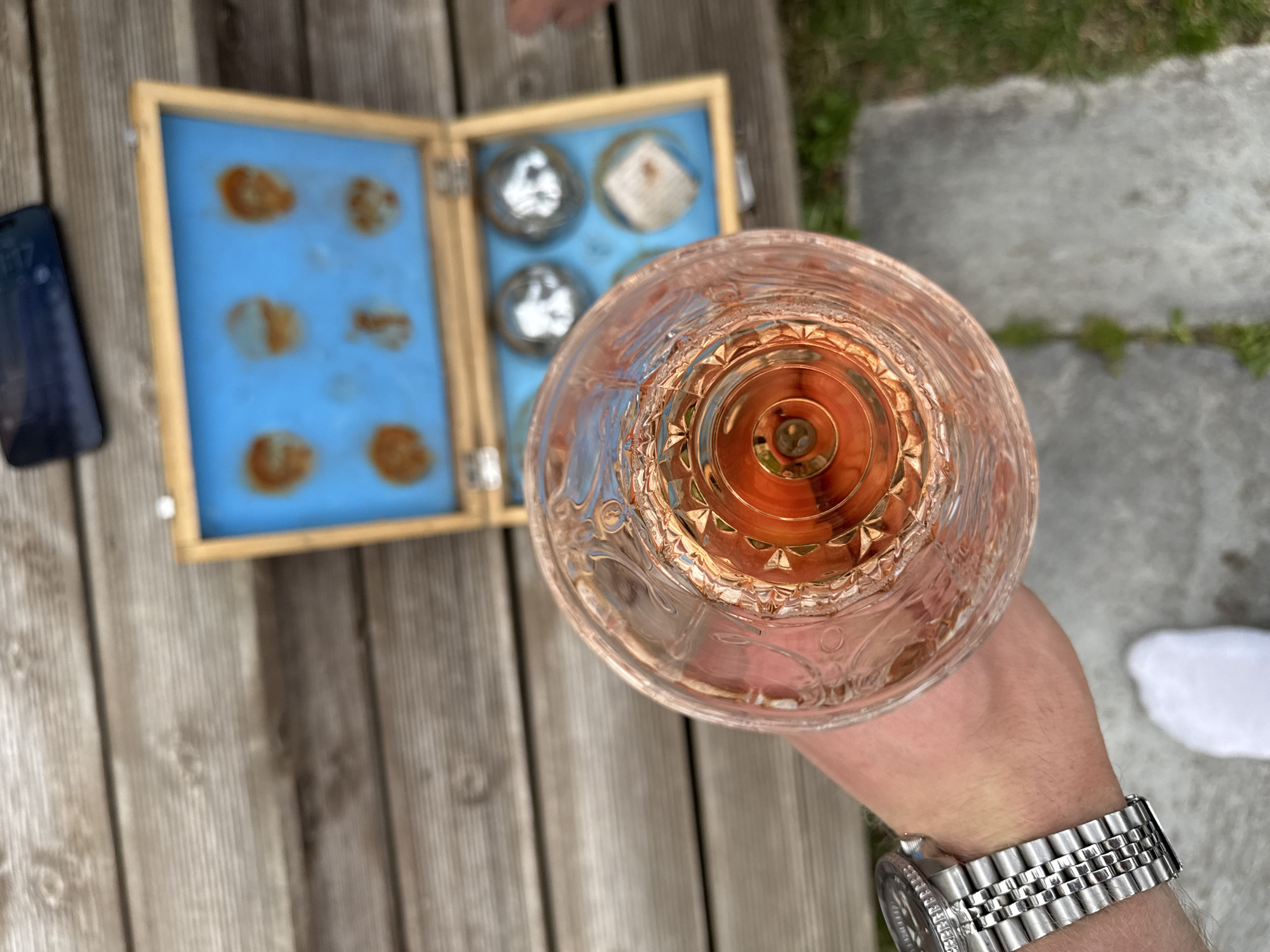 A close-up, top-down shot shows a glass of amber-colored liquid, possibly whiskey or brandy, held by a person wearing a stainless steel wristwatch. The glass is intricately patterned. In the blurred background, a wooden box with a blue interior is open, revealing what appear to be six spherical objects and a smaller square object within individual compartments. The scene is set on a wooden deck or table, with patches of grass and a gray, paved surface visible. The presence of a smartphone on the wooden surface suggests an outdoor setting. The location of Larkollen, Norway, is not explicitly depicted but is provided as context.