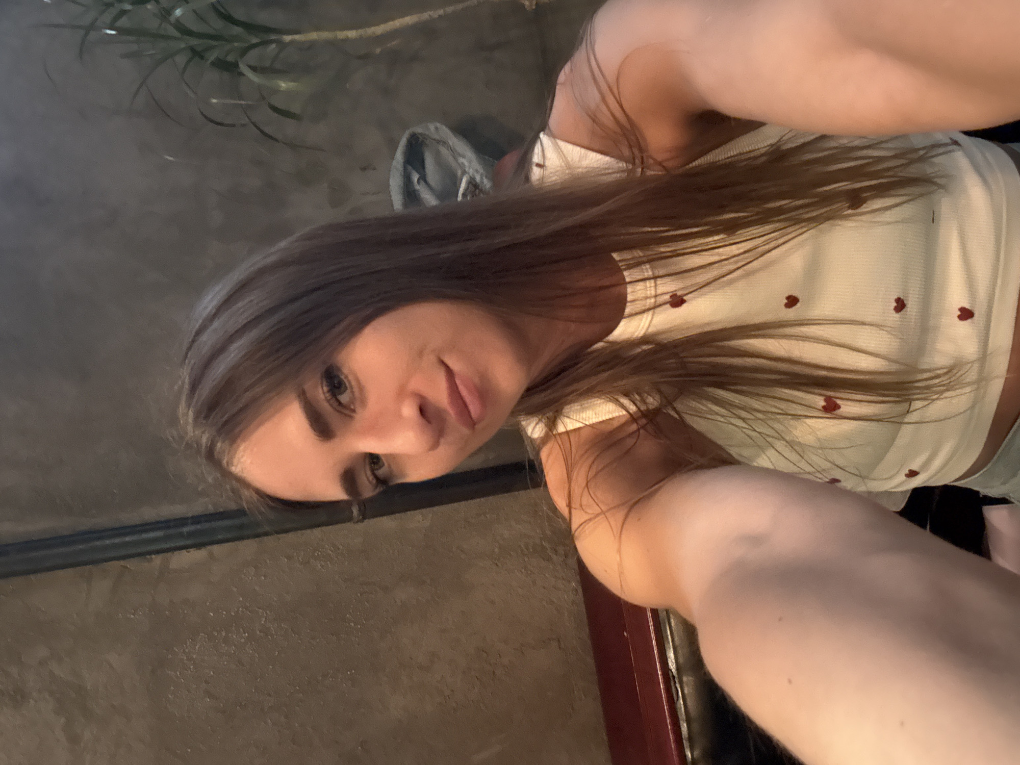 A young woman with long, brown hair and dark eyes is taking a selfie in what appears to be an indoor setting. She is wearing a white tank top adorned with small red hearts. Her face is turned towards the camera, and she has a slight smile. The background is dimly lit and textured, resembling a concrete wall. To the upper left, a plant with long, thin leaves is partially visible. To the lower left, there is a dark, metallic railing. The lighting suggests it might be evening or a dimly lit indoor space. The overall mood seems relaxed and casual. No visible text is present in the image.