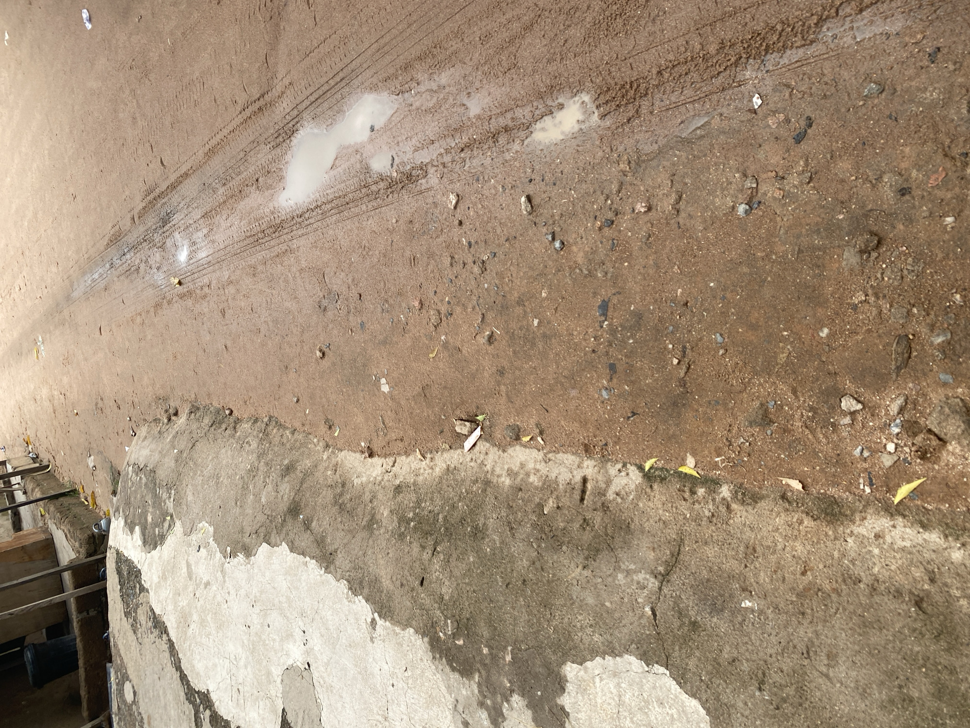 This is a close-up, eye-level shot of a dirt road in Jalingo, Nigeria. The road is unpaved and composed of reddish-brown soil, with visible tire tracks and several puddles of water indicating recent rain. Small stones and debris are scattered across the surface. To the left of the dirt road is a raised, concrete embankment. This embankment is weathered and cracked, with patches of lighter colored concrete and darker, wet areas. Some dried leaves are visible on both the concrete and the dirt. In the background, a row of structures, possibly shops or houses, are partially visible. These structures appear to be made of wood and concrete, with a dark-colored trash bin or container positioned near one of them. The overall impression is of a somewhat neglected urban or peri-urban environment.