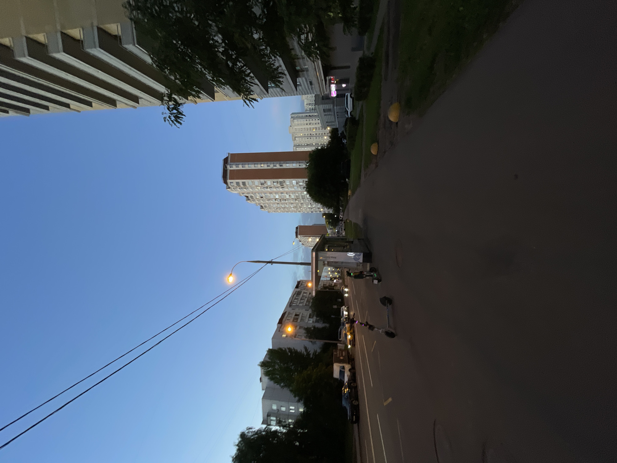 The image depicts a street scene in Moscow, Russia, during twilight. The sky is a clear, deep blue, transitioning from the day's light. On the right side of the frame, a tall, modern apartment building dominates the foreground, its balconies stark against the darkening sky. To its left, a pedestrian walkway lines the street, bordered by a patch of grass and a few spherical yellow bollards.
The street itself is a multi-lane road with cars driving and parked. A white van is visible, along with several passenger cars, their headlights on. On the sidewalk, near a bus stop shelter, two electric scooters are parked upright. The bus stop has a modern, glass-enclosed design. Across the street, a series of apartment buildings, typical of Russian urban architecture, are visible, some illuminated from within. Trees line the left side of the street, their foliage dark against the twilight sky. Overhead, power lines stretch across the frame, a common sight in urban environments. The overall impression is one of a residential area in a large city as evening descends.
