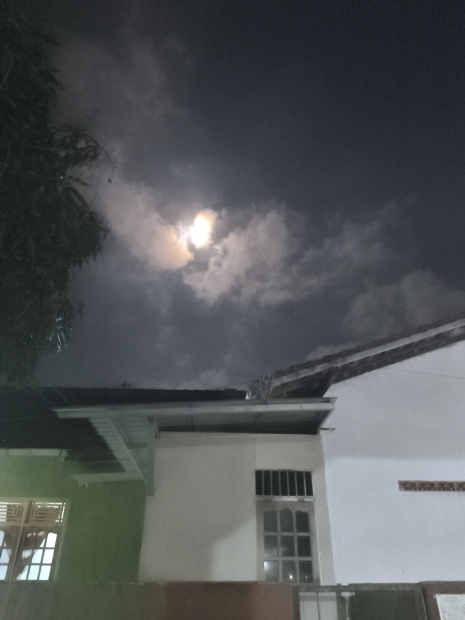 The image is a nighttime photograph taken in Kutoarjo, Indonesia, depicting a cloudy sky with a bright, partially obscured moon. In the foreground, the rooftops of houses are visible, constructed with a combination of dark tiles and lighter-colored eaves. One house has a white exterior wall and a window with a barred grille. Another house section to the left is painted green and features a window with multiple panes. A tree with dark foliage is present on the left side of the frame, partially obscuring the sky. The overall scene suggests a residential area at night, with the moon and clouds being the dominant natural elements in the sky.