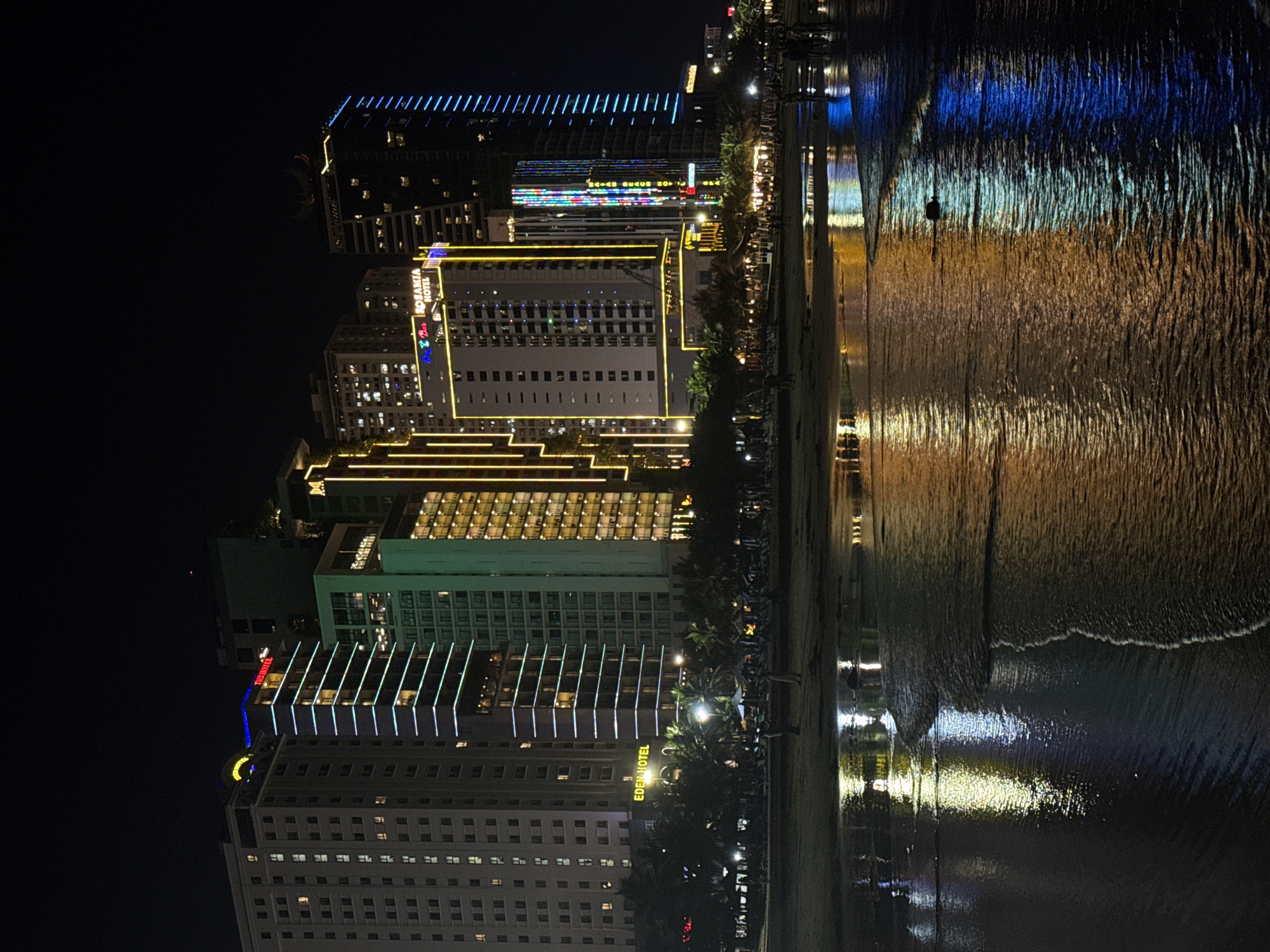 This is a nighttime, vertically oriented photograph of a beachfront urban landscape in Da Nang, Vietnam. The scene is dominated by several tall buildings, likely hotels, illuminated with a variety of colored lights. The buildings are positioned along a beachfront promenade, with a sandy beach and the ocean to their right.
On the left side of the image, we see a row of buildings. The furthest building has a prominent blue striped light pattern across its upper facade. Below this, other buildings are lit with yellow, white, and multi-colored lights, creating a vibrant cityscape. One building prominently displays the text "ROSAVIA HOTEL" in illuminated red and blue lettering, and another shows "EDEN HOTEL" in yellow. The architecture suggests modern high-rise structures.
The beach is visible to the right of the buildings, with the ocean beyond. The water reflects the lights from the buildings, creating shimmering patterns of blue, yellow, and white on its surface. Gentle waves are breaking on the shore. A few palm trees are visible along the promenade, their dark silhouettes contrasting with the illuminated buildings. The presence of beach chairs and umbrellas suggests this is a popular tourist area.
There are a few faint figures of people visible on the beach and promenade, appearing as dark silhouettes against the bright lights and reflections, indicating a relatively calm evening. The overall mood is lively and festive, characteristic of a tourist destination at night. The time of day is clearly night, with the dark sky providing a backdrop for the illuminated city and the glowing reflections on the water. The presence of the ocean and the style of the buildings are strong location cues for Da Nang, Vietnam.