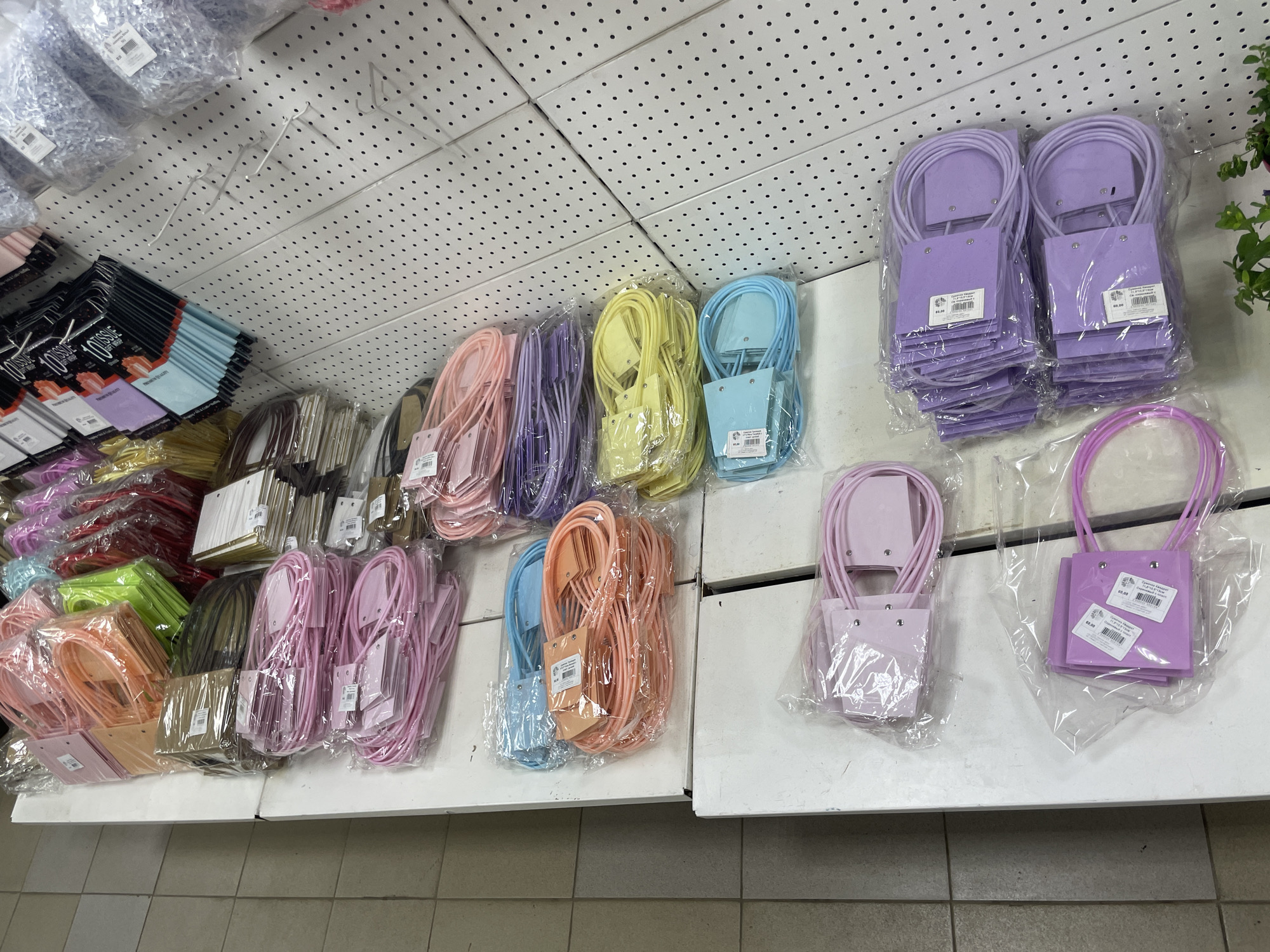The image shows a merchandise display in what appears to be a retail setting, specifically a store, in Unknown, Unknown. The display consists of multiple stacks of small, colorful shopping bags or pouches. These bags are neatly arranged in rows on a white shelf. The bags come in a variety of colors, including pink, peach, blue, yellow, and purple. Each bag is wrapped in clear plastic, indicating that they are new or for sale. The surface of the shelf has a tiled floor beneath it. The upper part of the background features a white, perforated panel ceiling, suggesting the location is an indoor commercial space. There are also some items on the shelves above, partially visible.