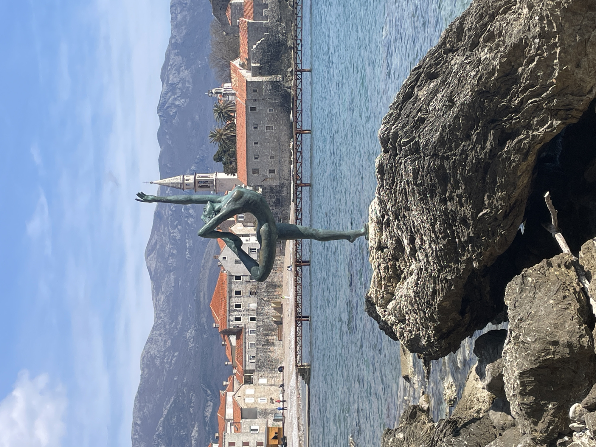 An outdoor, eye-level shot captures the "Dancer from Budva" statue in Budva, Montenegro, positioned on a large, textured rock in the foreground. The bronze statue, depicting a female figure in a dynamic ballet pose with arms extended upwards, stands on its left foot. Behind the statue, the calm, blue Adriatic Sea extends towards the Old Town of Budva. A wooden pier or boardwalk structure traverses the water, leading to a sandy beach.
The historic Old Town of Budva occupies the midground, characterized by stone buildings with orange-red tiled roofs and fortified walls. Prominent architectural features include a light-colored church tower with a conical spire on the left, and smaller domed structures. Sparse palm trees are visible within the townscape. A few indistinct human figures are visible on the sandy beach and near the Old Town buildings, some appearing to be walking or resting.
The background is dominated by large, rugged mountains under a clear blue sky with scattered white clouds. The overall scene depicts a sunny day on the Montenegrin coast.