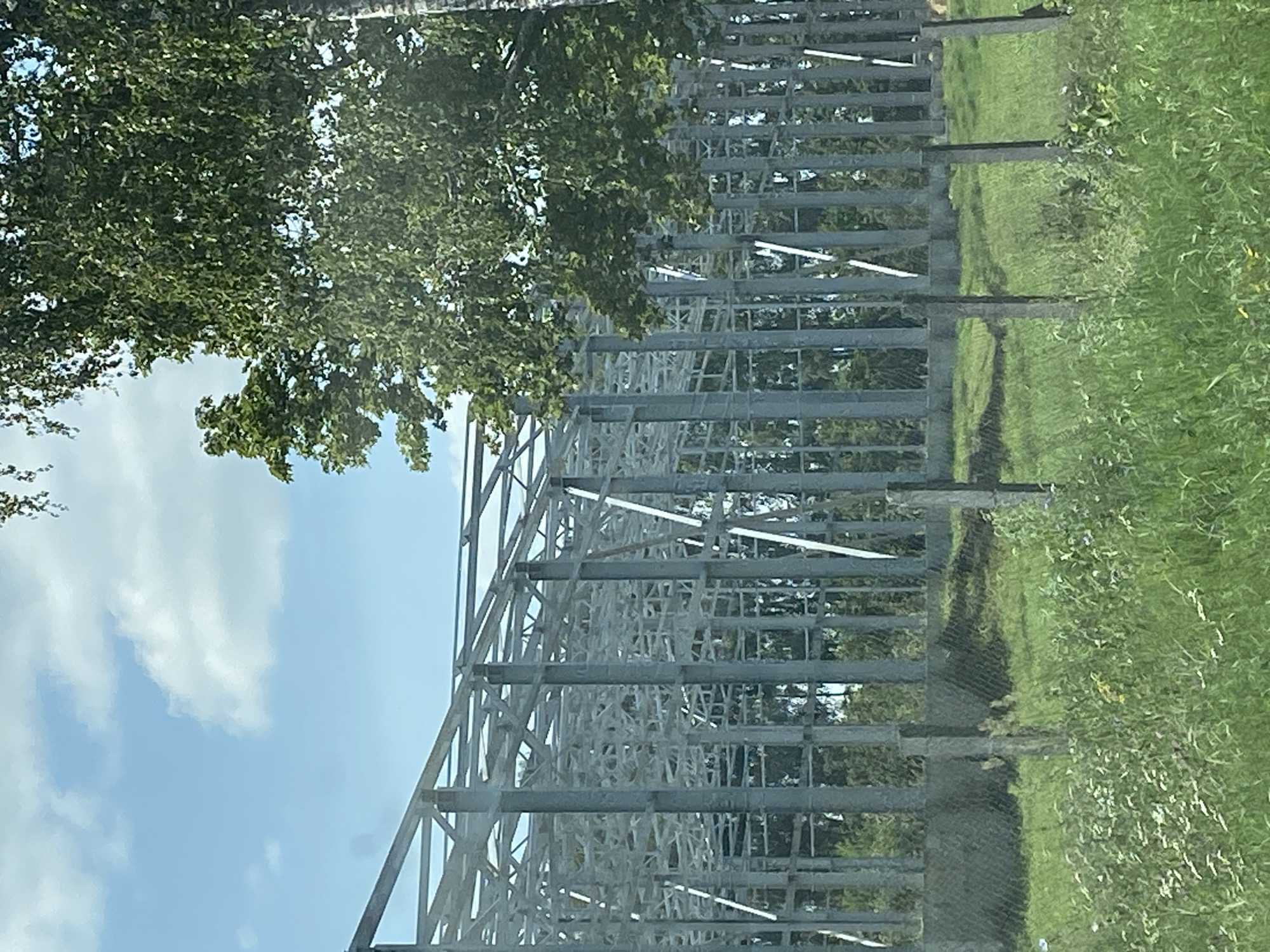 The image captures a construction site for a large, multi-story building, likely a warehouse or industrial facility, situated in the rural landscape of Воўчынскі сельскі Савет, Belarus. The skeletal structure of the building is dominated by a complex network of grey metal beams and columns, forming a vast, open framework. The construction is incomplete, with no visible walls or roofing, exposing the intricate internal bracing. Surrounding the site is a lush, green expanse of grassy fields and trees, with a prominent birch tree to the right of the frame providing a natural contrast to the industrial architecture. A chain-link fence runs along the perimeter of the construction area, with concrete posts interspersed with metal poles. The sky above is a clear blue with scattered white clouds, indicating fair weather. There are no people or vehicles visible in the scene, suggesting a temporary lull in the construction activity. The overall impression is one of ongoing development within a serene, natural environment.