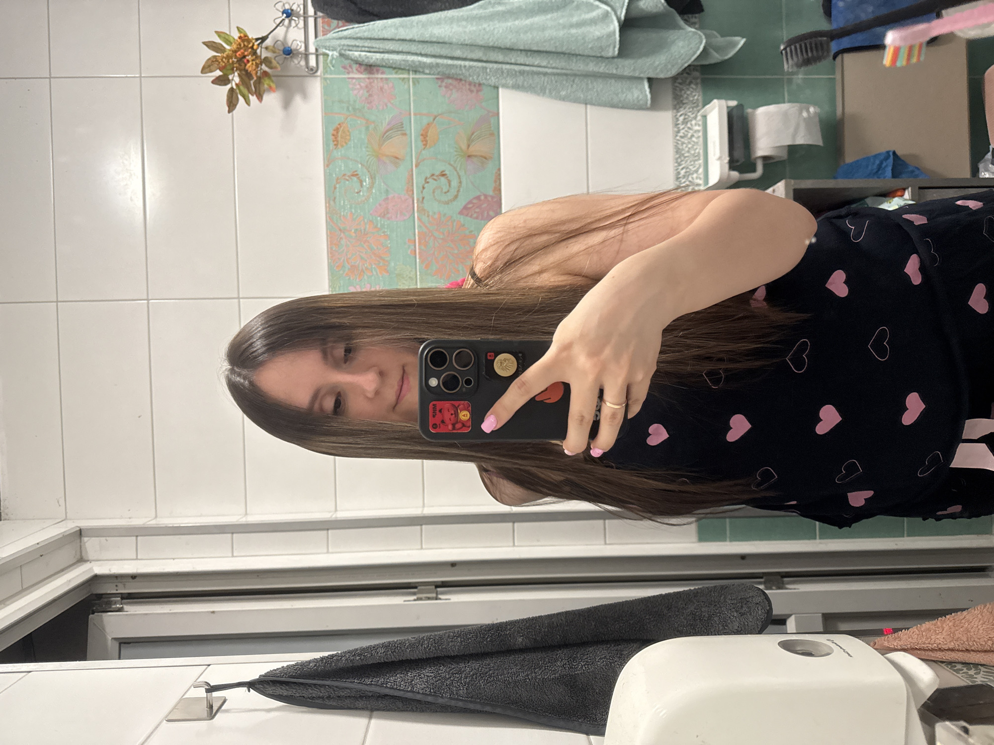 A young woman with long brown hair, wearing a black top adorned with pink hearts, takes a mirror selfie in a bathroom. Her right hand, adorned with a ring and a pink-painted fingernail, holds a black smartphone. The bathroom features white tiled walls, a green tiled backsplash with a floral pattern, and a silver-colored towel rack with a light blue towel hanging on it. To the left, a dark grey towel is draped over a hook, and a white dispenser is mounted on the wall. A pink towel is visible below the dispenser. In the background, a roll of toilet paper is mounted on the wall.