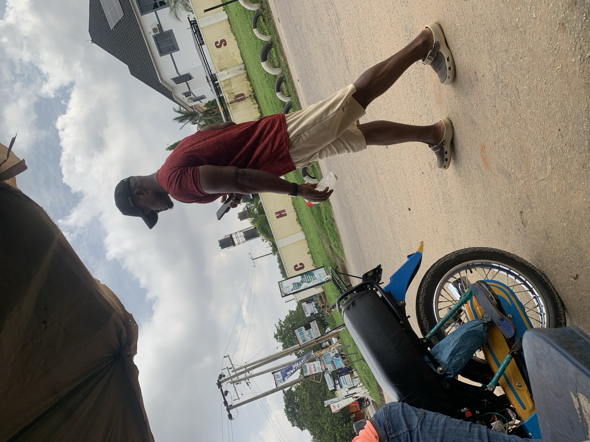 The image captures a man standing outdoors, holding a mobile phone and a plastic water bottle. He is wearing a red t-shirt, light-colored shorts, a black baseball cap, and tan clogs. The scene appears to be a street in Abuja, Nigeria, with a building featuring solar panels in the background. A blue and yellow tricycle is parked in the foreground. The weather seems cloudy, suggesting it could be daytime. The man's posture and the presence of the water bottle might indicate he has been exercising or is taking a break. Several billboards and utility poles are visible along the road, adding to the urban setting. One billboard displays the text "NADO" and another appears to have information about "FACIAL". The overall impression is one of a casual moment in a public space.