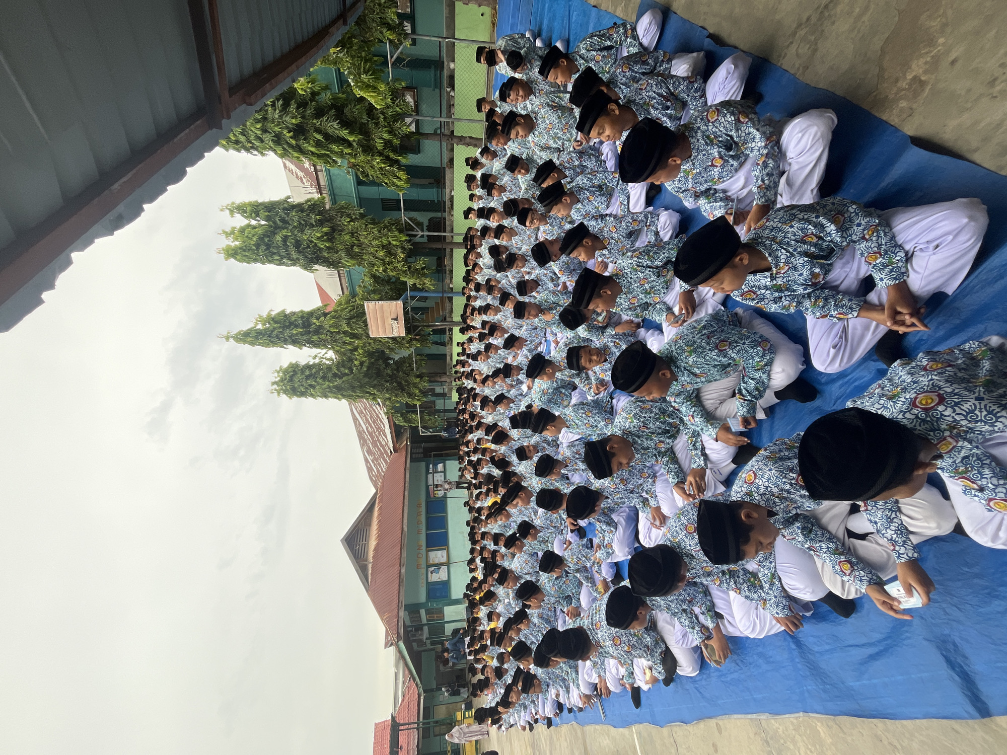 This image depicts a large group of young boys, likely students, seated on a blue tarp outdoors. They are all wearing similar traditional attire, consisting of white pants and patterned shirts with a blue and white design, and black skullcaps. The boys are arranged in rows, facing towards the left side of the frame, suggesting they are listening to or watching something. Some are holding small rectangular objects, possibly ID cards or papers.
The setting appears to be a schoolyard in Lhokseumawe, Indonesia. In the background, there are school buildings with green walls and red tiled roofs. Tall, slender trees with dense green foliage line the area between the students and the buildings. The sky is overcast with a light gray hue. The overall atmosphere suggests an organized gathering, possibly a school assembly or event.