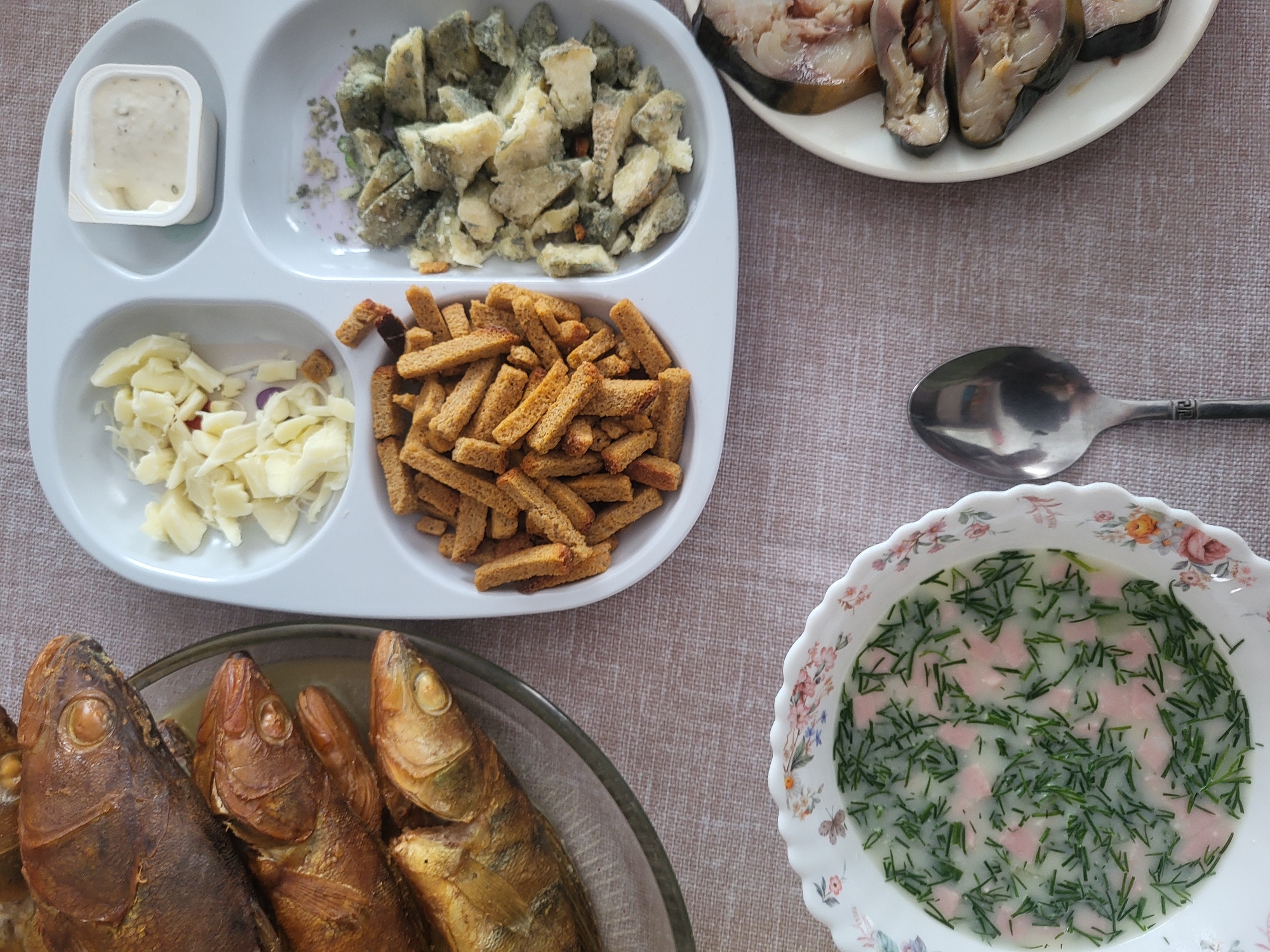 The image presents a table laden with food. In the lower left, there is a glass plate filled with four smoked fish. Above it, a white partitioned tray holds a small container of white sauce, chunks of a grayish-blue cheese, small white cubes (possibly cheese or garlic), and croutons. Above the tray, a plate contains slices of smoked mackerel. A spoon rests near the center of the image, and in the lower right is a bowl of soup with pink cubes and chopped green herbs. The setting is in Зипуново, Russia.