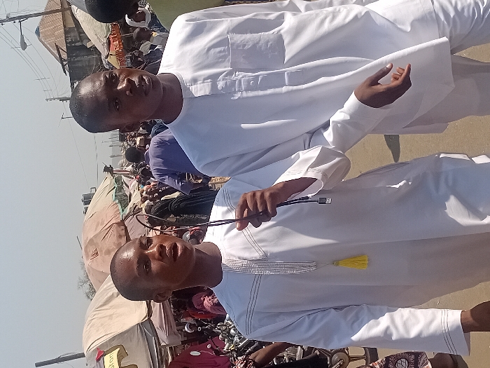 Two men wearing white traditional attire are posing outdoors. The man in the foreground holds a cable with a yellow tassel attached to his garment. Buildings and tents dot the background with people milling about. The picture is a close-up shot, showcasing the subjects' faces and upper bodies.