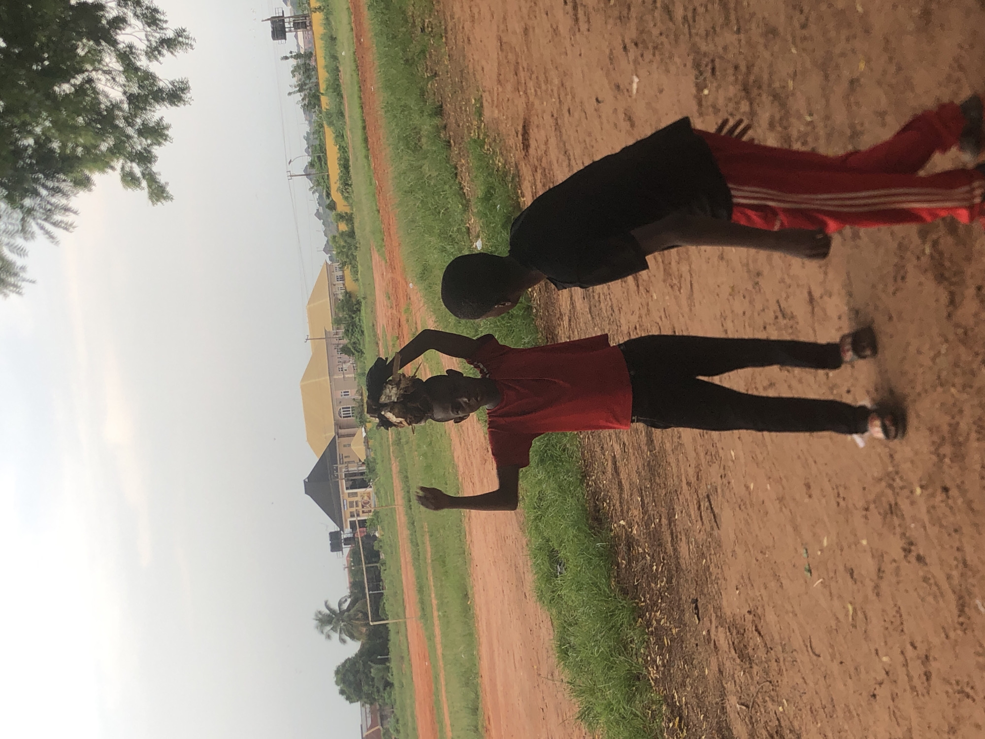 Two young individuals are captured outdoors. One person wears a red shirt and dark trousers and is holding something above their head. The other wears a black shirt and red tracksuit bottoms with white stripes. They are on a dirt patch adjacent to a grassy area, possibly a sports field in Ukwu-Abwa, Nigeria. Buildings are visible in the background. A tree is visible in the upper left corner.