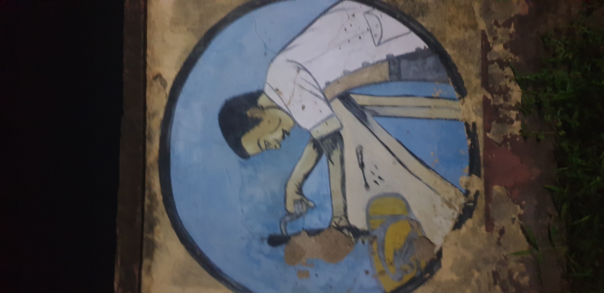 The image shows a circular mural on a distressed wall. The mural depicts a person, likely a barber, in the act of cutting hair. The barber is shown from the chest up, wearing a white shirt with a pocket. They are holding a pair of scissors and are focused on cutting the hair of another individual whose head is obscured by the barber's action. The background of the mural is a solid light blue. The wall itself is textured and shows signs of age and damage, with peeling paint and rough patches, particularly around the base where greenery is visible. The overall scene is dimly lit, suggesting it might be evening or night. The mural is located in Sabon Tasha, Nigeria, an area known for its vibrant street art.