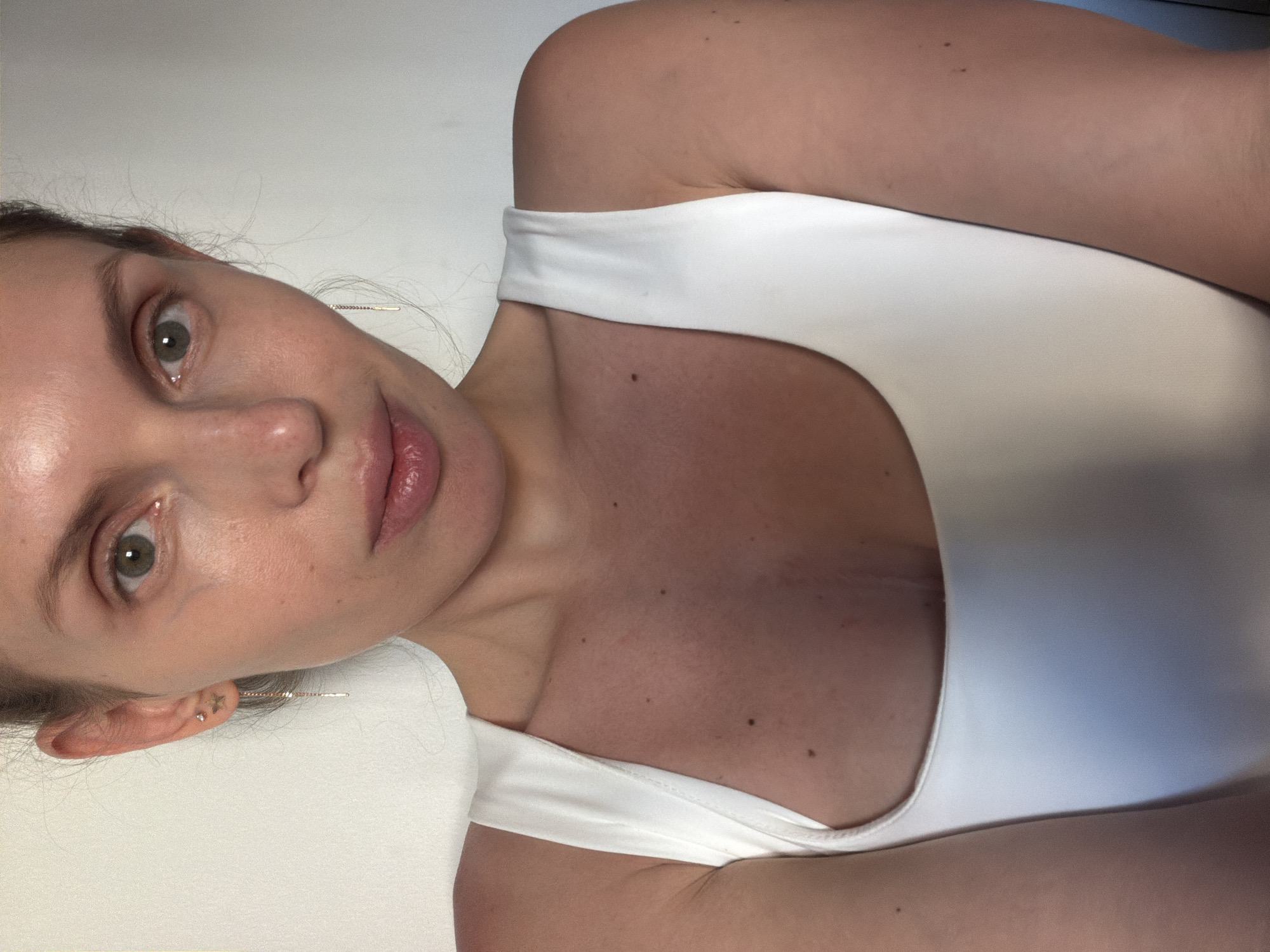 A woman with fair skin, light eyes, and pink lips is wearing a white top. She is positioned with her face in the left side of the frame, and her body at an angle. A hair pin is visible in her hair behind her ear. The environment is a flat white surface, with a hint of blue in the upper right corner. There is no indication of Budva, Montenegro in the image.