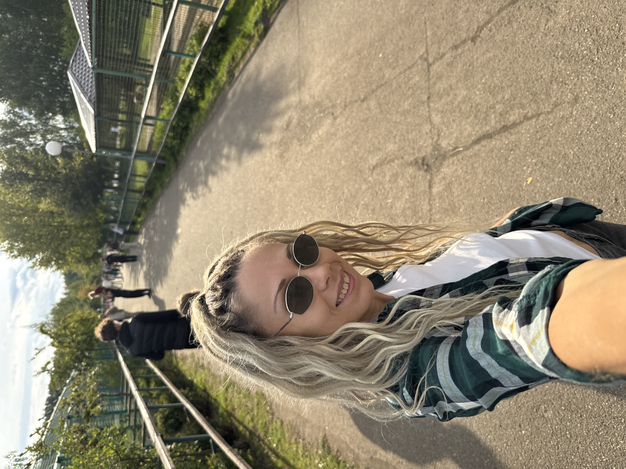 A woman with blonde, curly hair and sunglasses takes a selfie outdoors. She is wearing a green plaid shirt over a white t-shirt. She is smiling at the camera. Behind her, a paved path winds through a park-like setting with trees and a fence. Other people are visible in the background, walking along the path. The location is the city of Ignatovo, Russia.
