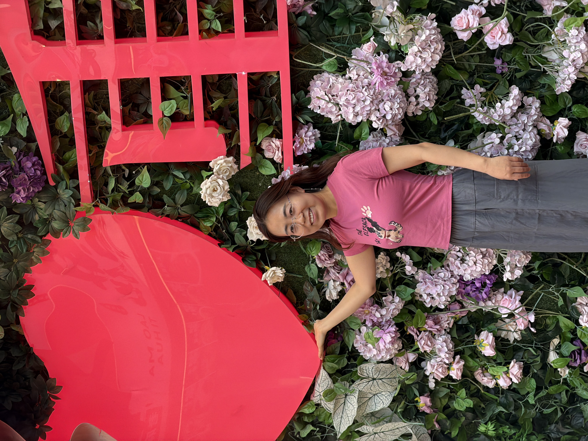 A woman with glasses and a pink t-shirt stands in front of a floral wall display. She is smiling at the camera. The wall is decorated with a large red heart on the left and red Chinese characters on the right. The backdrop is a dense arrangement of artificial greenery, pink and white flowers, and some larger white patterned leaves. The woman's left arm is extended, and her hand is resting on the bottom edge of the red heart. The setting appears to be outdoors, possibly a public space or event in Yuzhong District, China.