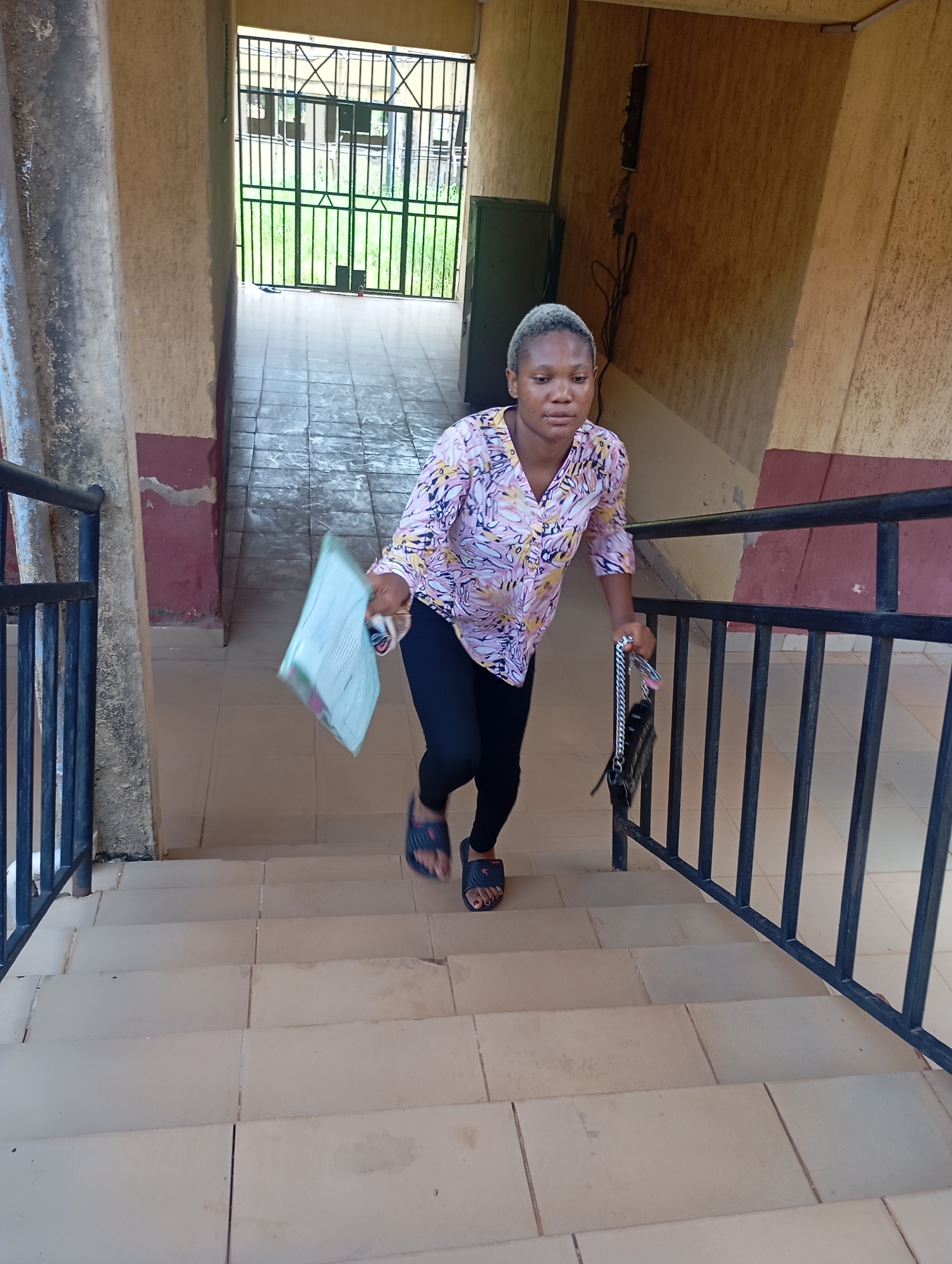 A young woman ascends a tiled staircase in what appears to be a semi-enclosed passage or stairwell in Ukwu-Abwa, Nigeria. She is positioned slightly to the right of the center, captured mid-stride as she walks upwards. Her short, light-colored or blonde hair frames a neutral expression. She wears a long-sleeved, patterned shirt featuring abstract pink, yellow, and white designs, paired with black leggings and dark blue slide sandals, revealing red painted toenails on one foot.
In her left hand, she carries a light green document or booklet, and in her right, a black purse with a silver chain strap. A black metal railing lines the right side of the staircase, providing support. The stairs are made of light-colored tiles, some showing minor wear and discoloration. The walls of the passage are painted in two tones: a darker reddish-brown along the lower section and a lighter, sandy-beige above, exhibiting signs of age and wear.
In the background, at the top of the stairs, a dark green metal gate opens to an area with green grass, suggesting an outdoor space or courtyard beyond. Overhead, some electrical wiring and a small utility box are visible on the upper wall. The lighting indicates it is daytime, providing clear illumination for the scene. No discernible text is visible in the image.