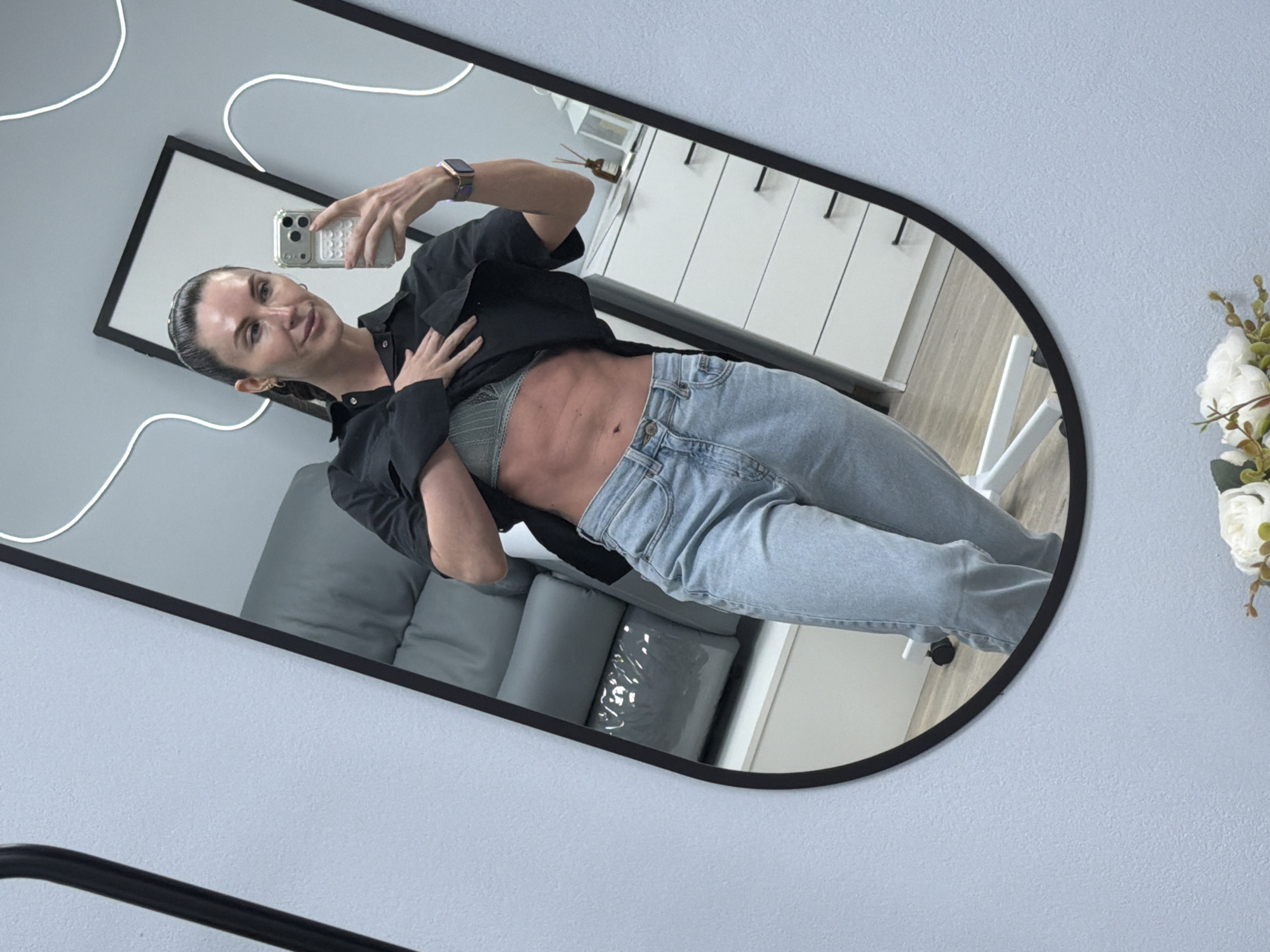 A woman is reflected in a black-framed oval mirror. She is holding a phone in her right hand, taking a selfie. She is wearing light blue jeans, a grey-green bra, and an open black shirt. The reflection shows a grey couch with pillows behind her and a white cabinet to her left. A white decorative light fixture is visible on the wall behind the mirror. A floral arrangement is to the right of the mirror. The wall behind the mirror appears to be a light blue. The city location, Perm, Russia, is not verifiable from the image.