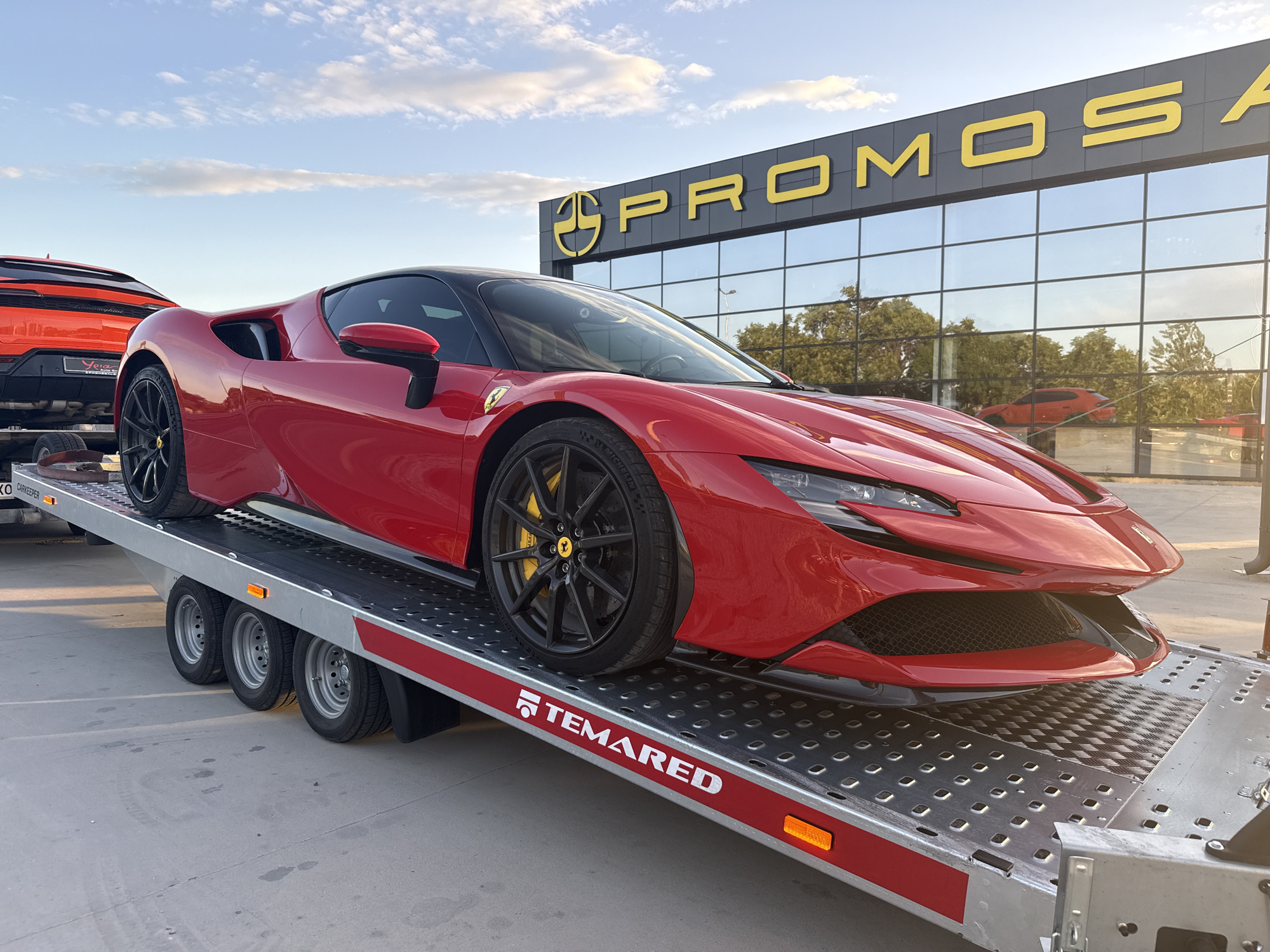 A red Ferrari SF90 Stradale sits on a gray Temared car trailer parked on a paved surface. Behind the Ferrari, and partially visible, is the rear of an orange Lamborghini Urus. The backdrop features a modern building with large, mirrored windows and the company name "PROMOSA" in large, yellow letters atop a dark gray background. The reflection in the windows shows trees and another vehicle on the trailer. The scene is likely taken in Plovdiv, Bulgaria, based on the company name and registered number plates of the trailer visible (KO). The sky is a mix of blue and light clouds, suggesting either early morning or late afternoon lighting. The cars appear to be newly delivered.