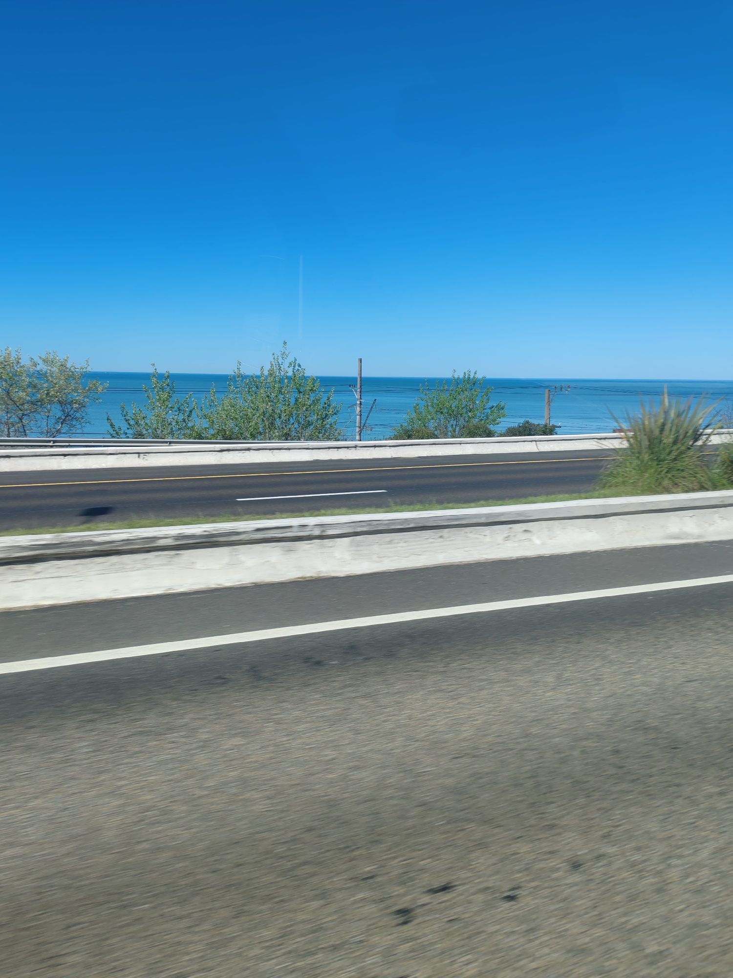 The image shows a road with a view of a body of water and a clear blue sky. Trees and vegetation line the side of the road, near the water's edge. There are utility poles visible in the background. The road itself has white and yellow lines marking lanes. The location appears to be near a coastline, with the road providing a view of the water. The city and specific location are unknown based on the provided image.