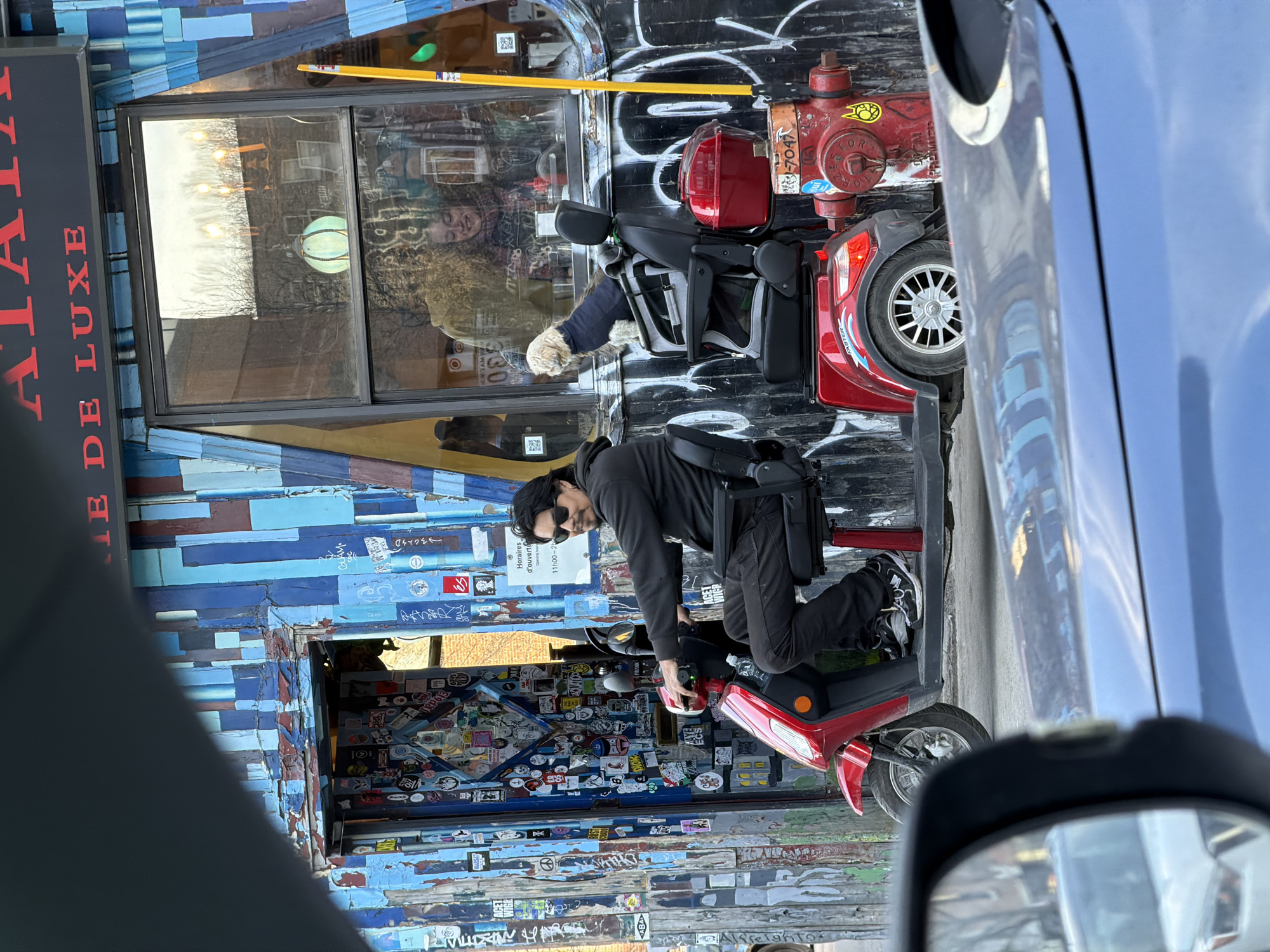 Here is a description of the image:
The image depicts a man operating a red mobility scooter in front of a building in Montreal, Canada. The building's facade is decorated with various stickers and the sign "Atelier De Luxe." The man is wearing a dark-colored outfit and sunglasses. In the background, there is a fire hydrant near the building. A dog in a mobility scooter can be seen in the building's window. The photograph appears to be taken from inside a vehicle, showing parts of the car's interior like the rearview mirror and the side window frame. The entire image is oriented vertically.