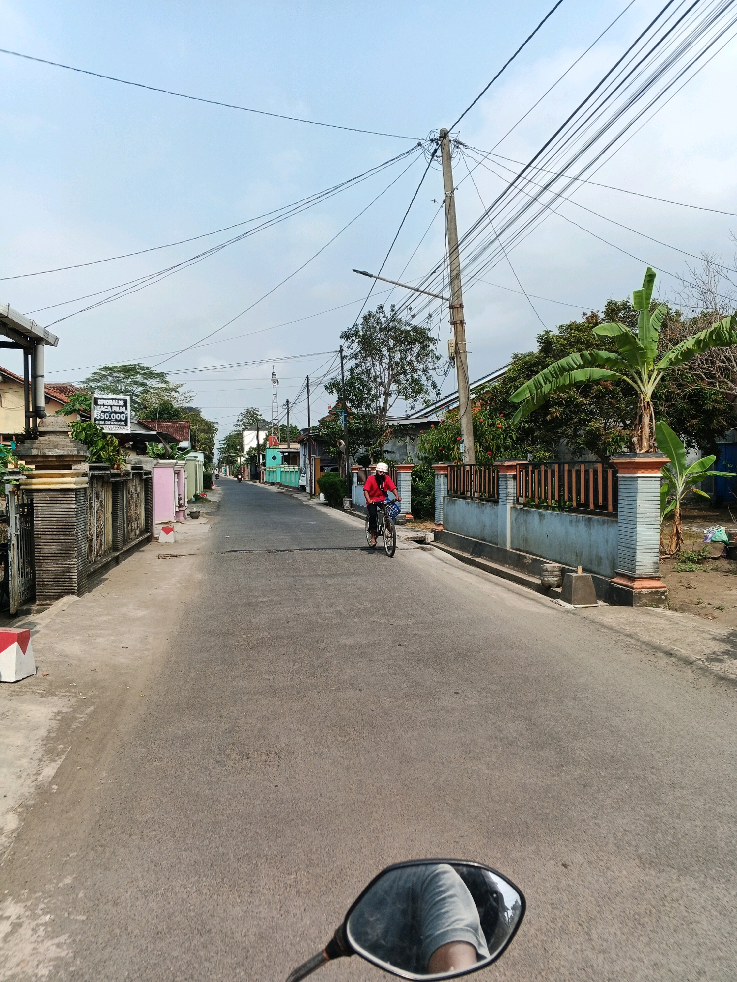 This image depicts a street scene in Jangrana, Indonesia. In the foreground, the mirror of a motorcycle reflects a person's arm and torso wearing a grey t-shirt. The motorcycle is moving down an asphalt road lined with houses and fences. In the middle ground, a person wearing a red shirt and a white helmet is riding a bicycle. The person is carrying a blue bag in the basket of the bicycle. To the left of the road, there is a fence with decorative pillars and a sign that reads "SPESIALIS KACA FILM 350.000 BISA DITANGGIL". To the right of the road, there is a banana tree and more fencing. The sky is clear with some scattered clouds. Power lines are visible crisscrossing the sky. The overall environment appears to be a residential area with a mix of modern and traditional architecture.
