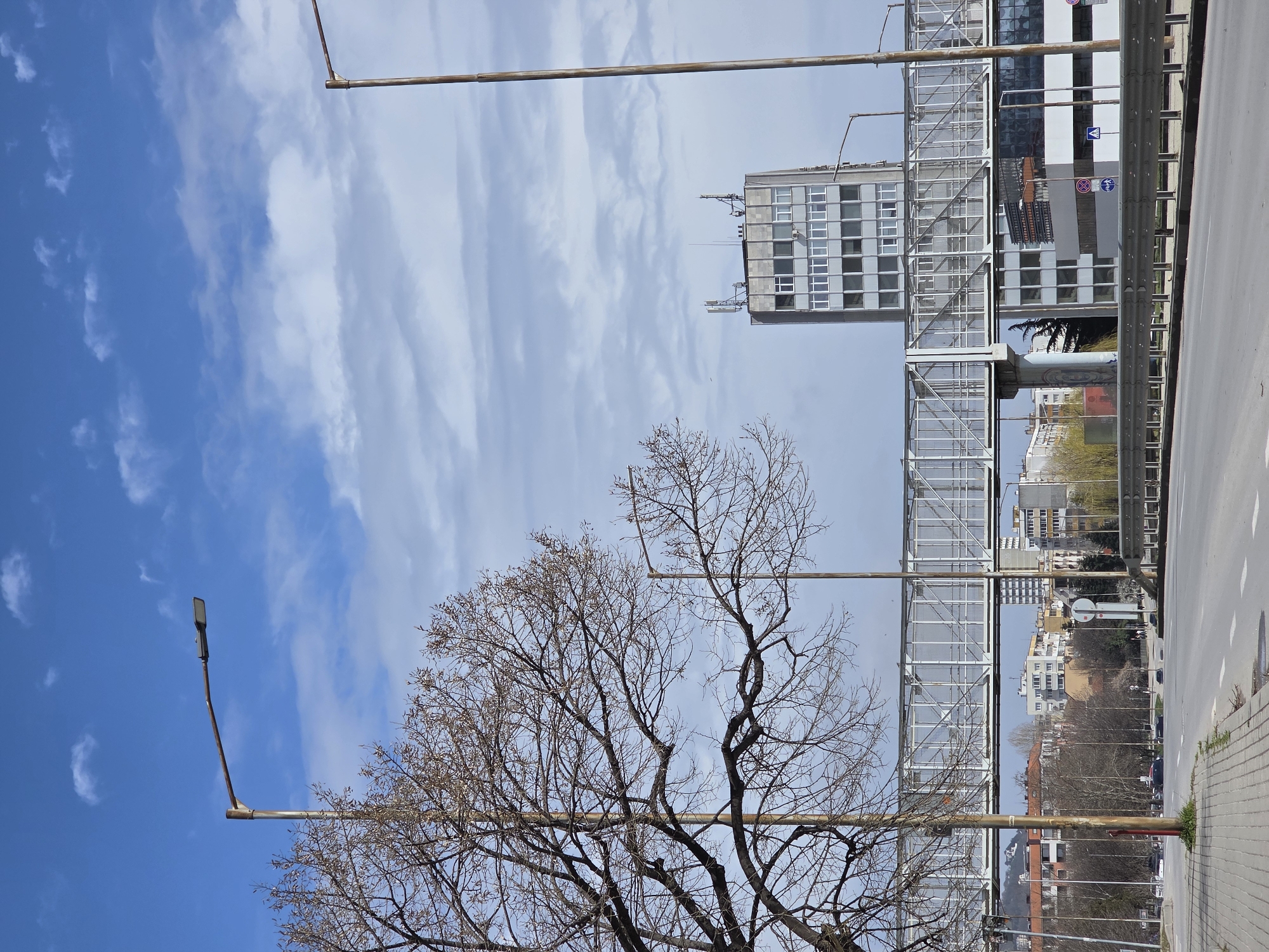 The image captures a daytime urban street scene in Varna, Bulgaria, specifically along Vladislav Varnenchik Boulevard near the Grand Mall, characterized by a prominent pedestrian overpass and the "Varna Tower" building. The sky is blue with scattered white stratocumulus clouds.
In the foreground, a light-grey paved sidewalk occupies the left, bordering a multi-lane asphalt road marked with white lines. A metal guardrail runs along the right edge of the road. Two tall, weathered metal streetlamp posts, fitted with modern LED luminaires, stand on opposing sides of the road. A large, bare deciduous tree with an extensive network of branches partially obscures the upper left quadrant.
The midground is dominated by a substantial grey metal pedestrian overpass, featuring a lattice-work design and transparent side panels, spanning horizontally across the image. It is supported by concrete pillars. Below the overpass, road signs are visible, including a red-and-blue "No stopping or standing" sign and a blue "Pedestrian crossing" sign.
In the background, to the right, a tall, multi-story, rectangular grey concrete building, known as "Varna Tower," stands prominently, featuring multiple rows of windows and topped with communication antennas. Additional buildings of varying heights and residential structures are visible in the distance, set against a backdrop of distant, indistinct hills. The scene appears to lack active vehicle traffic or pedestrians, depicting a quiet street environment.