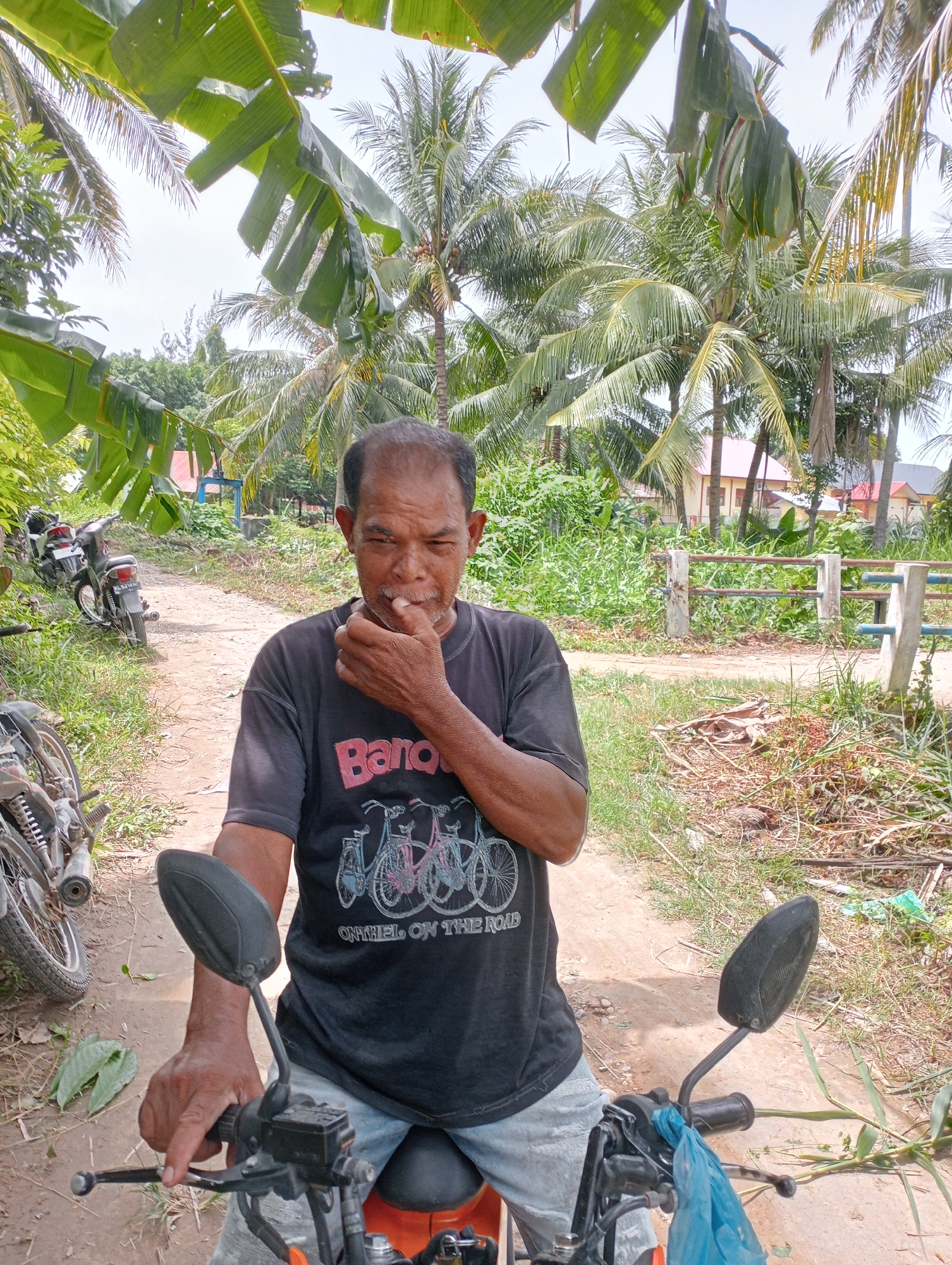 An adult male sits on a parked orange motorcycle on a dirt road, surrounded by lush tropical vegetation. The man wears a black t-shirt with a bicycle print and light-colored pants. He raises his right hand to his mouth. Behind the man and the motorcycles, a rustic fence line is visible, beyond which is dense greenery and a few houses with red roofs. Palm trees and banana plants loom in the background, filling the frame.