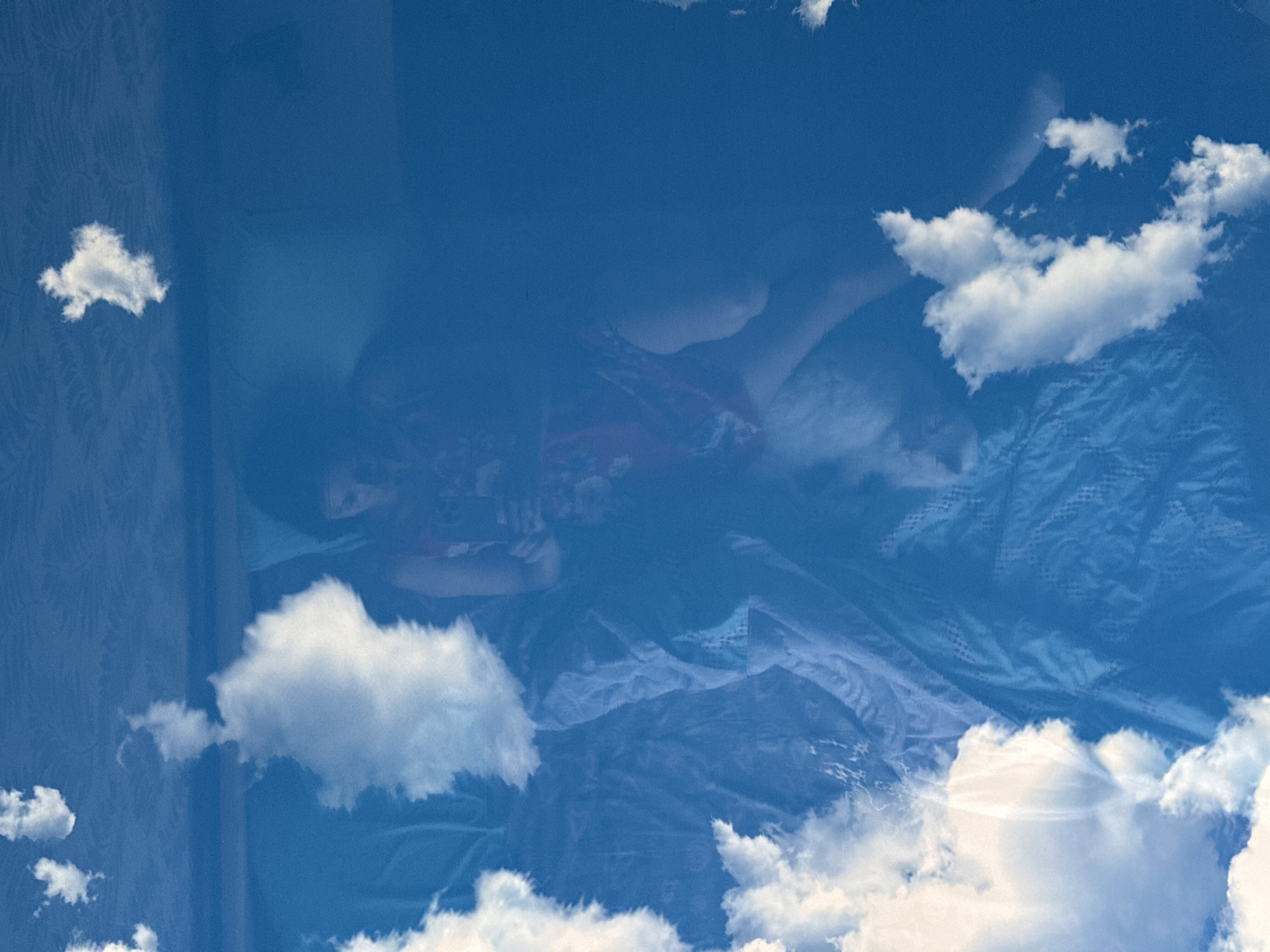 A person is lying in a bed, overlaid with an image of clouds in a blue sky. The location is indoors, implied by the presence of furniture and wallpaper on the left side of the image. The person appears to be resting or sleeping. The overall impression is one of surrealism or dreamlike quality due to the superimposed sky. No location is explicitly specified in the city of Unknown, Unknown.